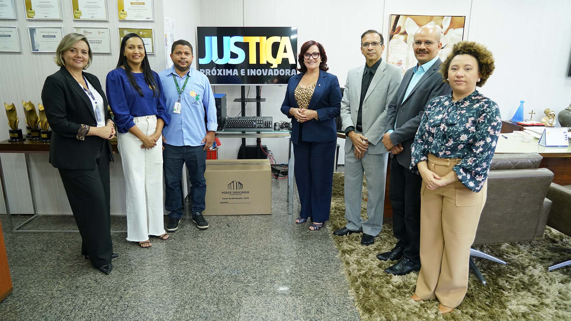 Homens e mulheres em pé em frente a uma tv com dizeres sobre a Justiça durante entrega de computadores