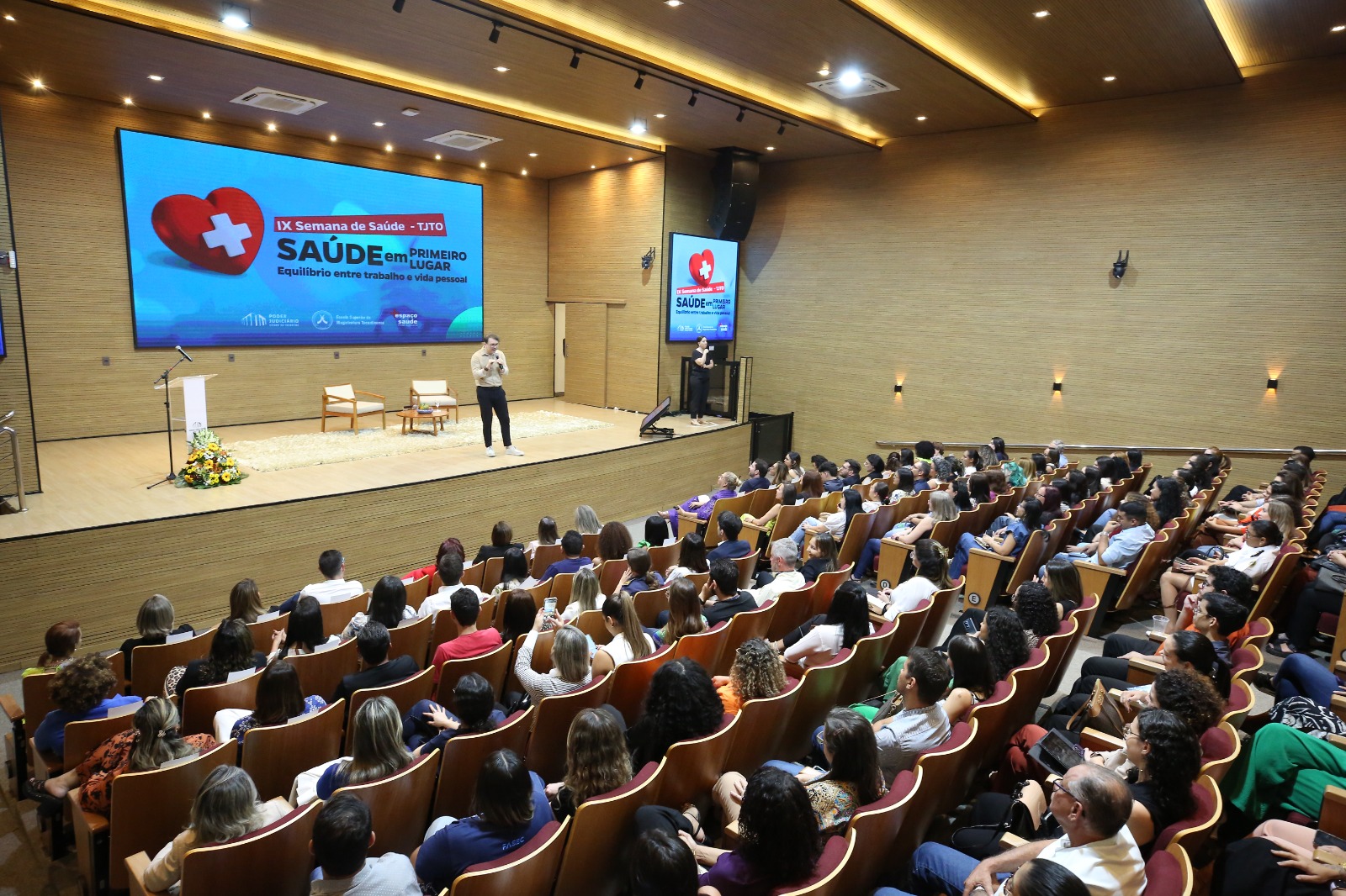 Auditório lotado de magistrados e servidores sentados e no palco o palestrante em pé, de frente, falando ao microfone. Atrás um monitor grande com a imagem da Semana de Saúde em fundo azum e um coração vermelho