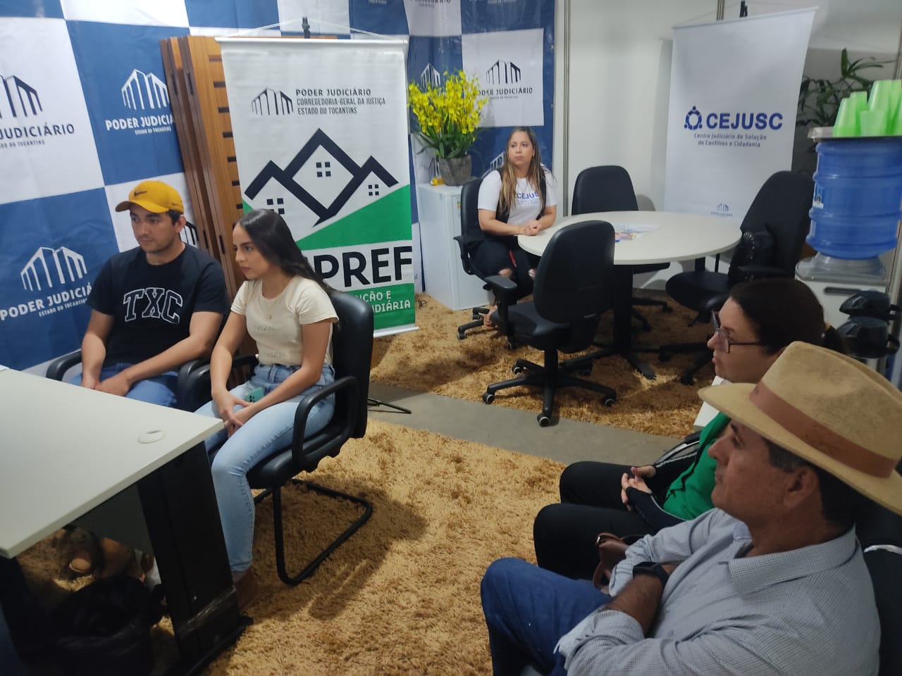 Homens e mulheres sentados em um estande do Judiciário enquanto aguardam atendimento. No chão, tapete marrom, ao fundo, mesa redonda com uma mulher de cabelos claros e longos e camiseta branca sentada. Nas paredes, adesivo azul e branco do Judiciário e um banner branco e verde com dizeres do Núcleo de Regularização Fundiária