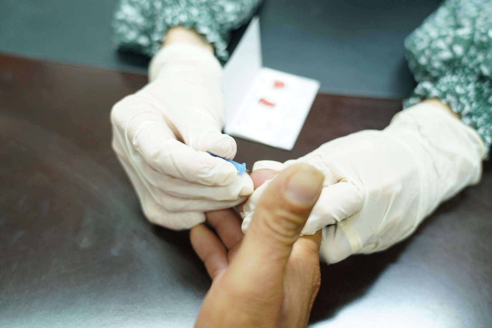 imagem de duas mãos com luvas coletando o sangue de um dos dedos de um mão masculina; na foto também aparece um papel com marcas de sangue coletado.