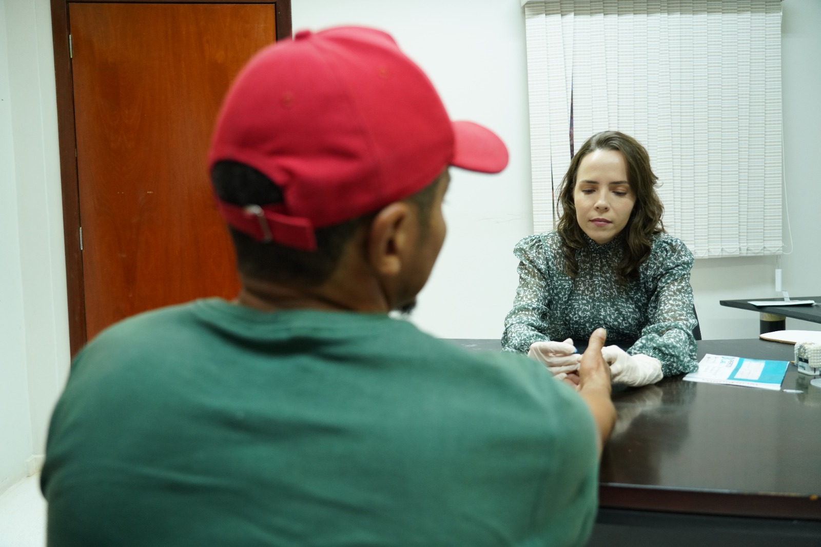 Homem de boné vermelho e camiseta verde, sentado de costas coletando sangue para exame de DNA; à frente uma servidora da Justiça sentada coletando o exame