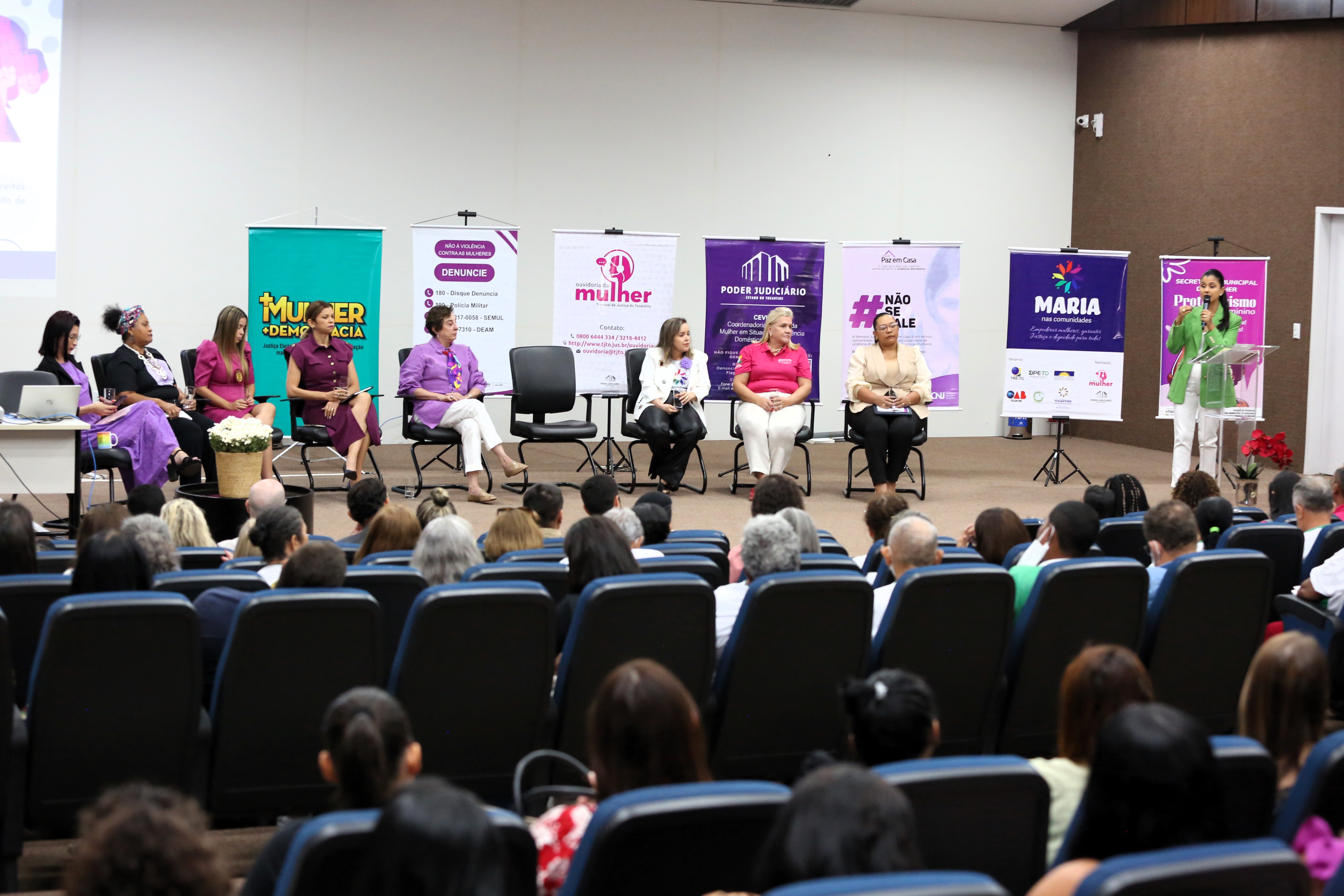 auditório lotado; no pauco nove mulheres, sendo uma desembargadora e duas juízas, participam de uma roda de conversa; oito estão sentadas e uma está à direita, falando ao microfone; ao fundo vários banners de programas do Judiciário de proteção à mulher