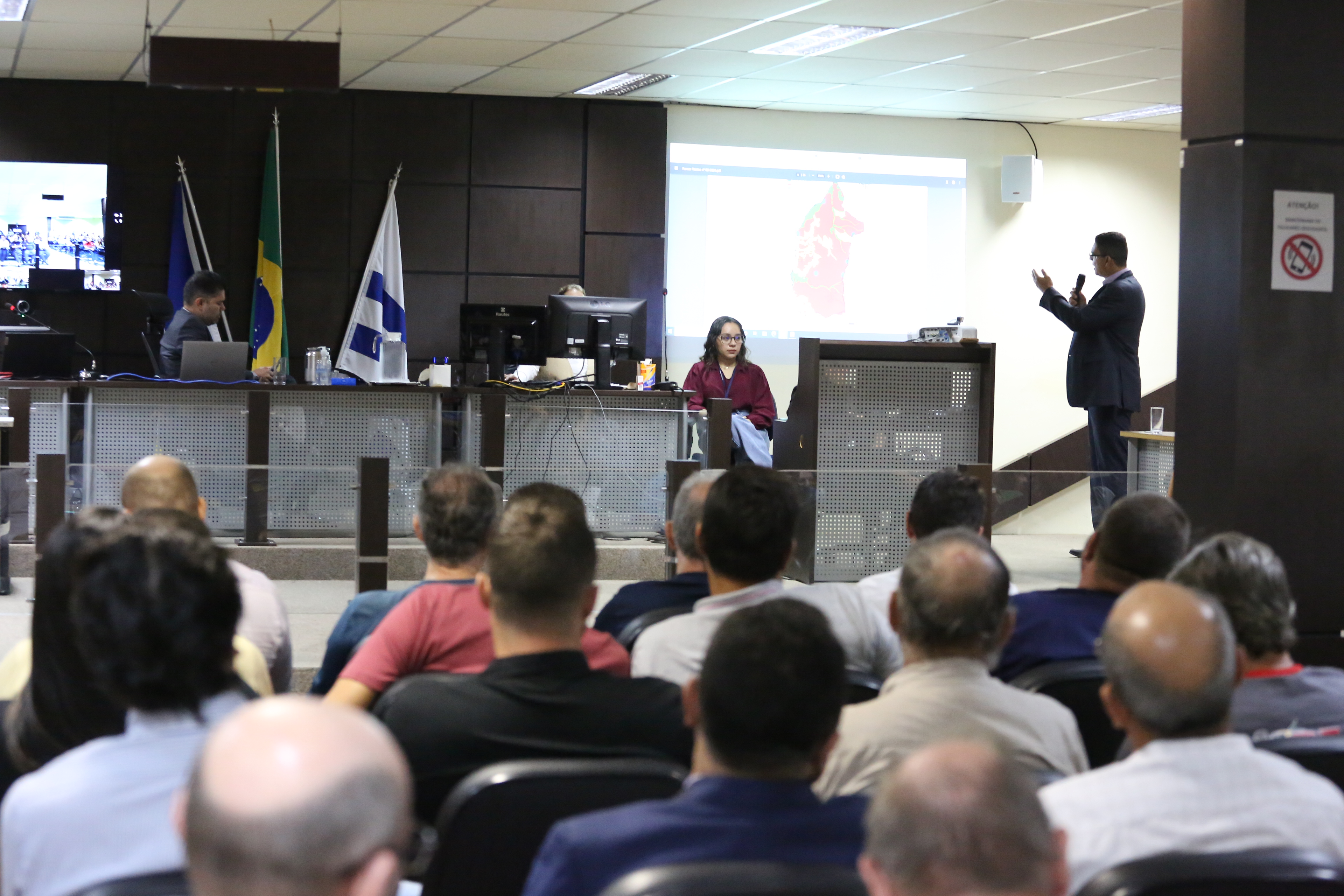 fotografia em formato de paisagem mostra pessoas de costas, sentadas assistindo à uma explanação de homem que usa terno cinza escuro e está de pé falando ao microfone e apontando uma projeção de slides na parede. ao lado esquedo da imagem o magistrado está sentado de perfil e obsevando