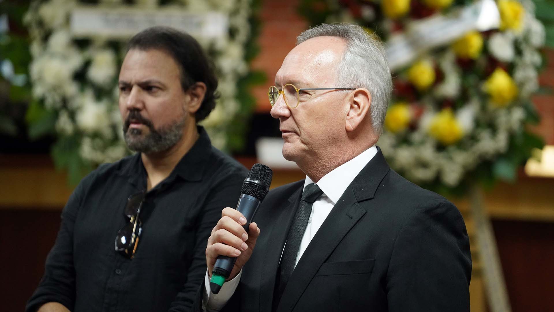 Fotografia colorida que mostra um homem branco grisalho, usando óculos e segurando um microfone. Ele usa terno preto, ao fundo vê-se outro homem moreno, de barba, e duas coroas de flores