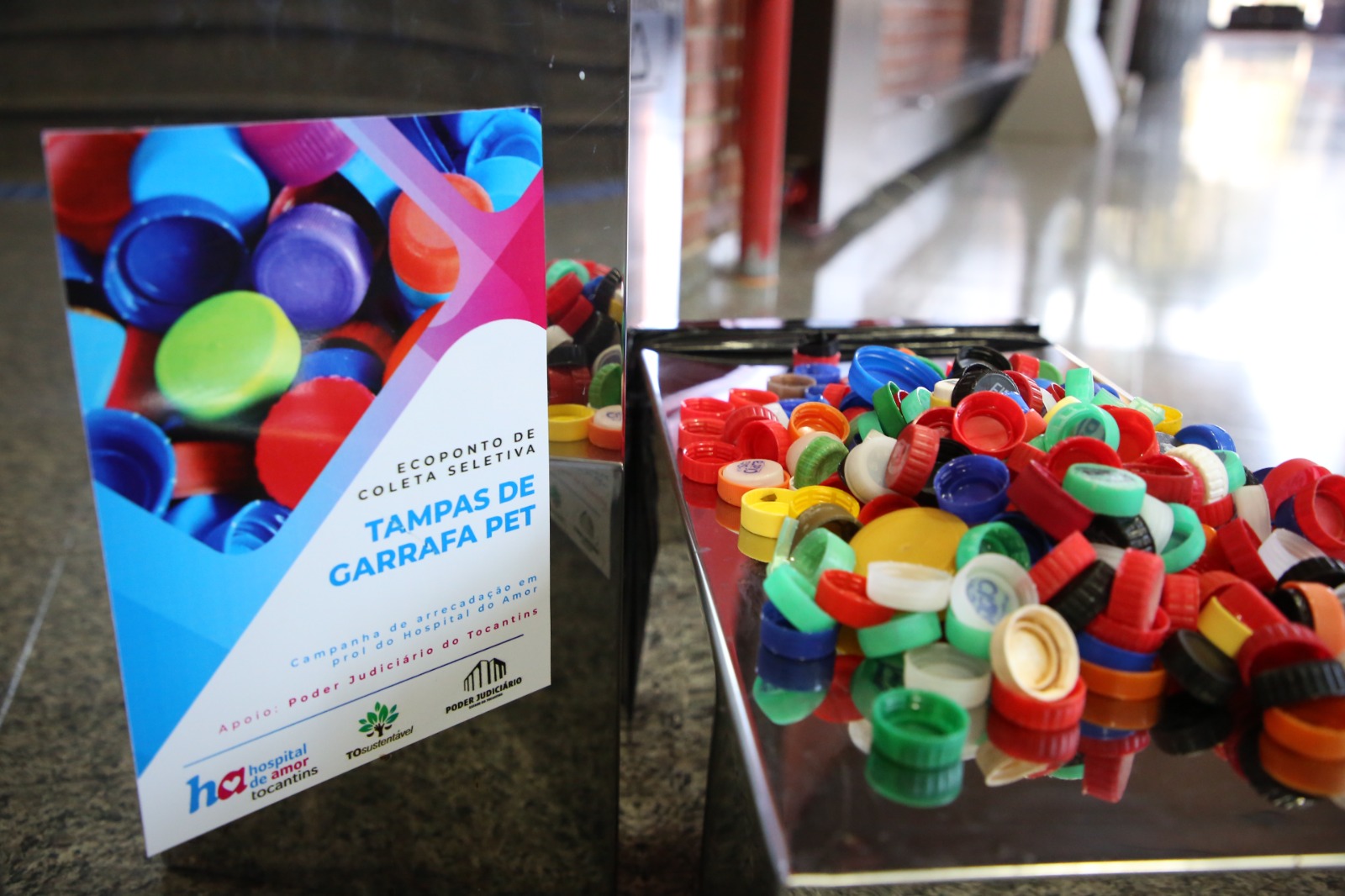 Imagem mostra cartaz sobre campanha de arrecadação de tampinhas plásticas. Ao lado, tampas arrecadadas