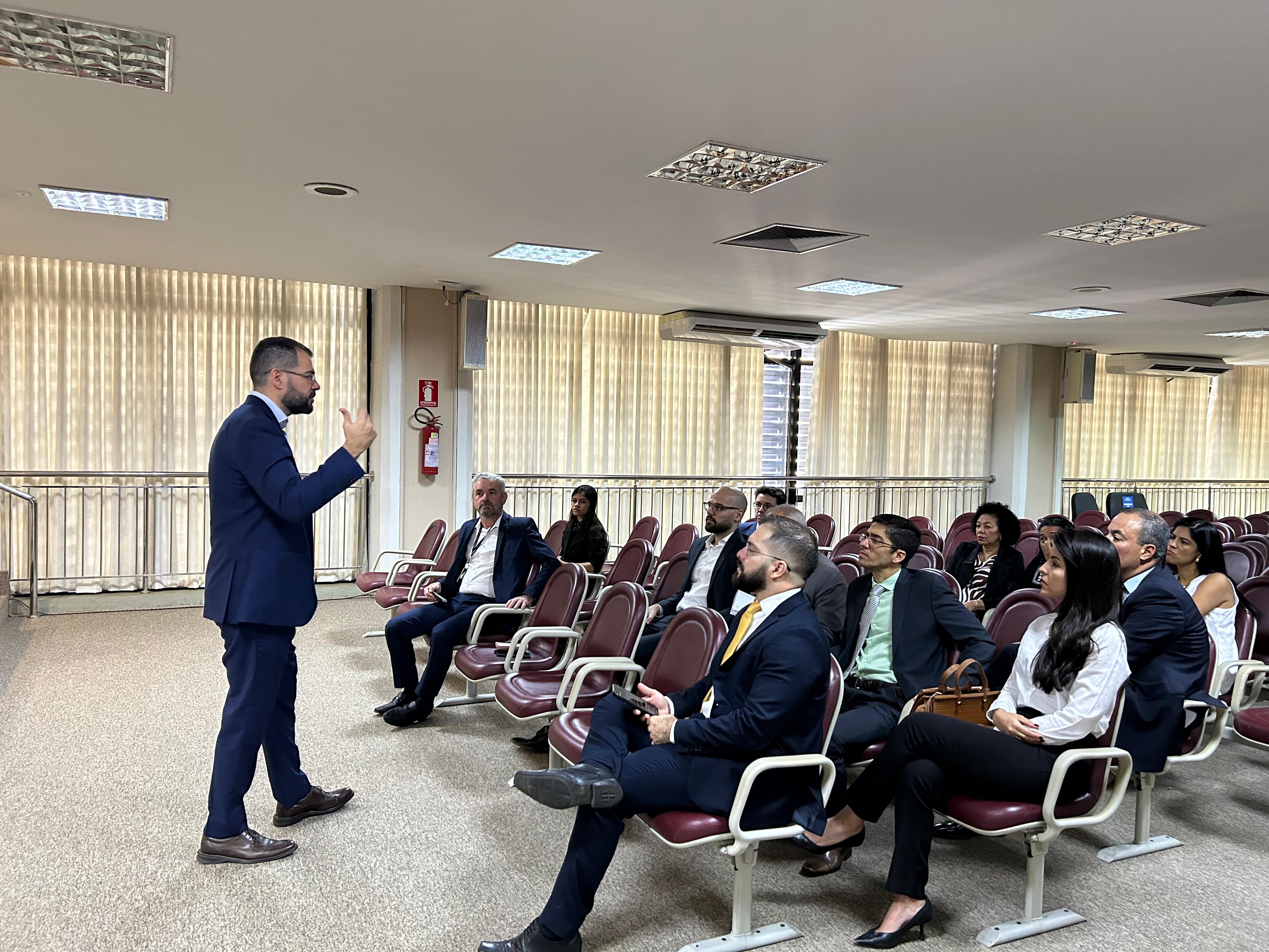 Homem pardo, magro, cabelos e barba rentes, veste terno azul escuro e fala pra público de homens e mulheres sentados em cadeiras de cor vinho de auditório com carpete bege no piso e paredes claras, com persianas beges