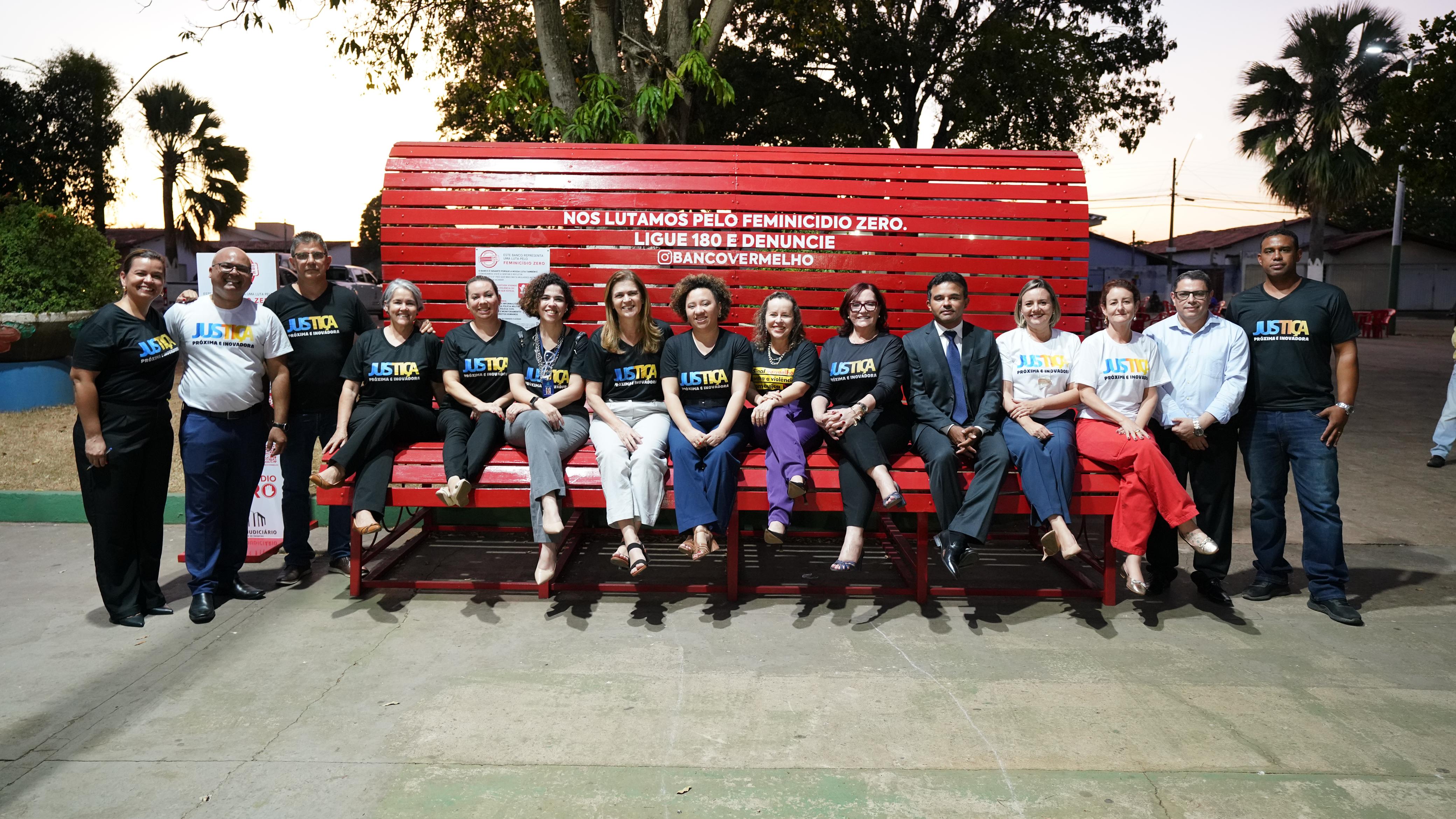 Grupo de 14  pessoas, algumas em pé e outras sentas em um banco vermelho gigante.