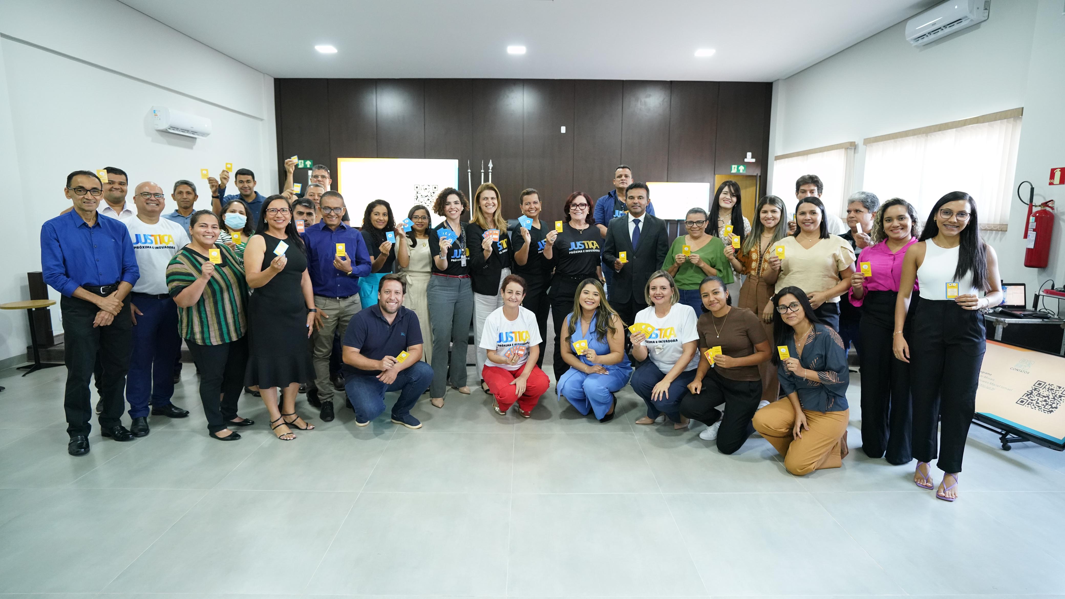 Homens e mulheres dentro de sala de paredes laterais brancas e parede de fundo de madeira escura. Eles posam para foto segurando um cartão amarelo