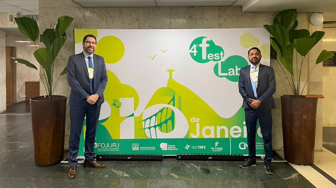 foto colorida horizontal mostra dois homens vestindo terno, eles sorriem e estão entre dois grandes vasos de planta e em frente a um extenso painel que anuncia o 4 festlab