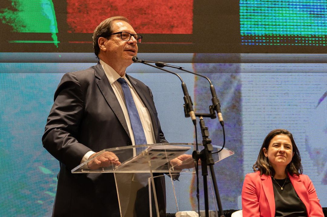 foto colorida horizontal mostra homem branco discursando ao microfone. Ele usa óculos com moldira preta e vesta terno azul marinho, com camisa branca e gravata azul
