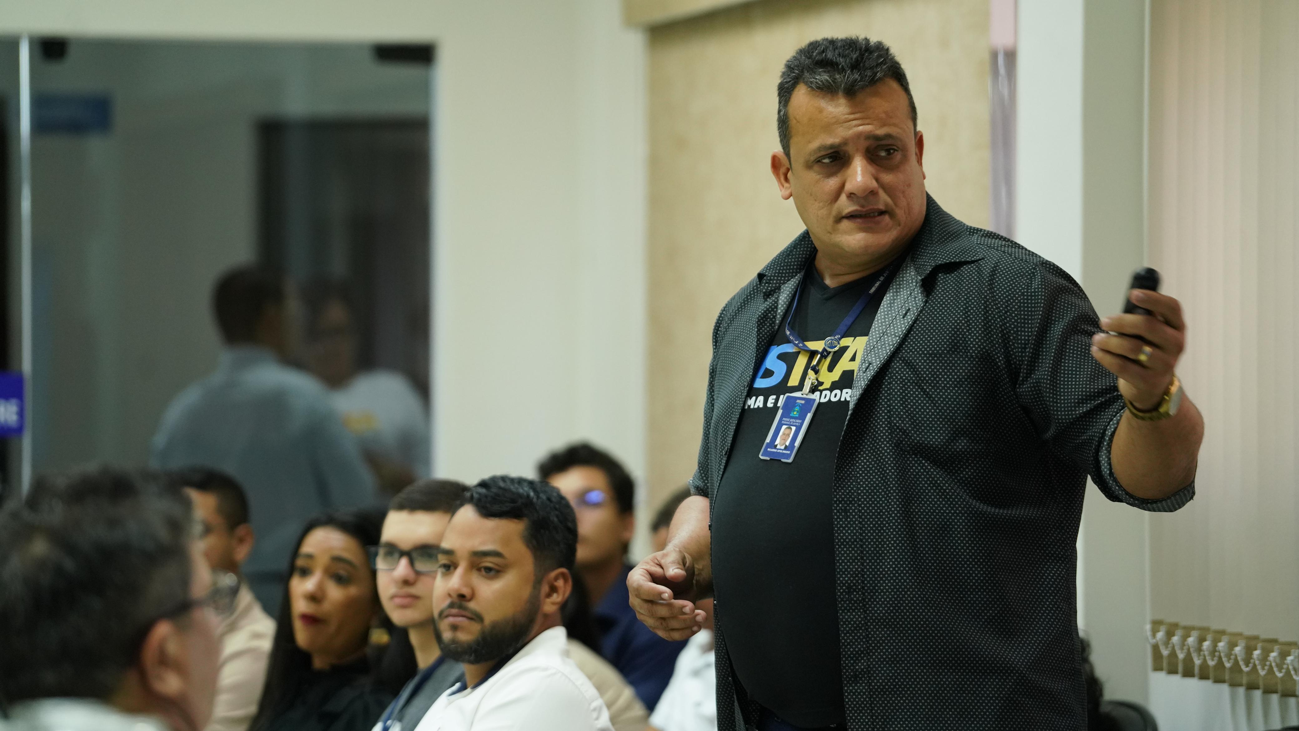 Imagem colorida que mostra um homem usando preto, com a camisa escrito Justiça mais próxima e apontando com um controle para frente. Ao fundo há várias pessoas 