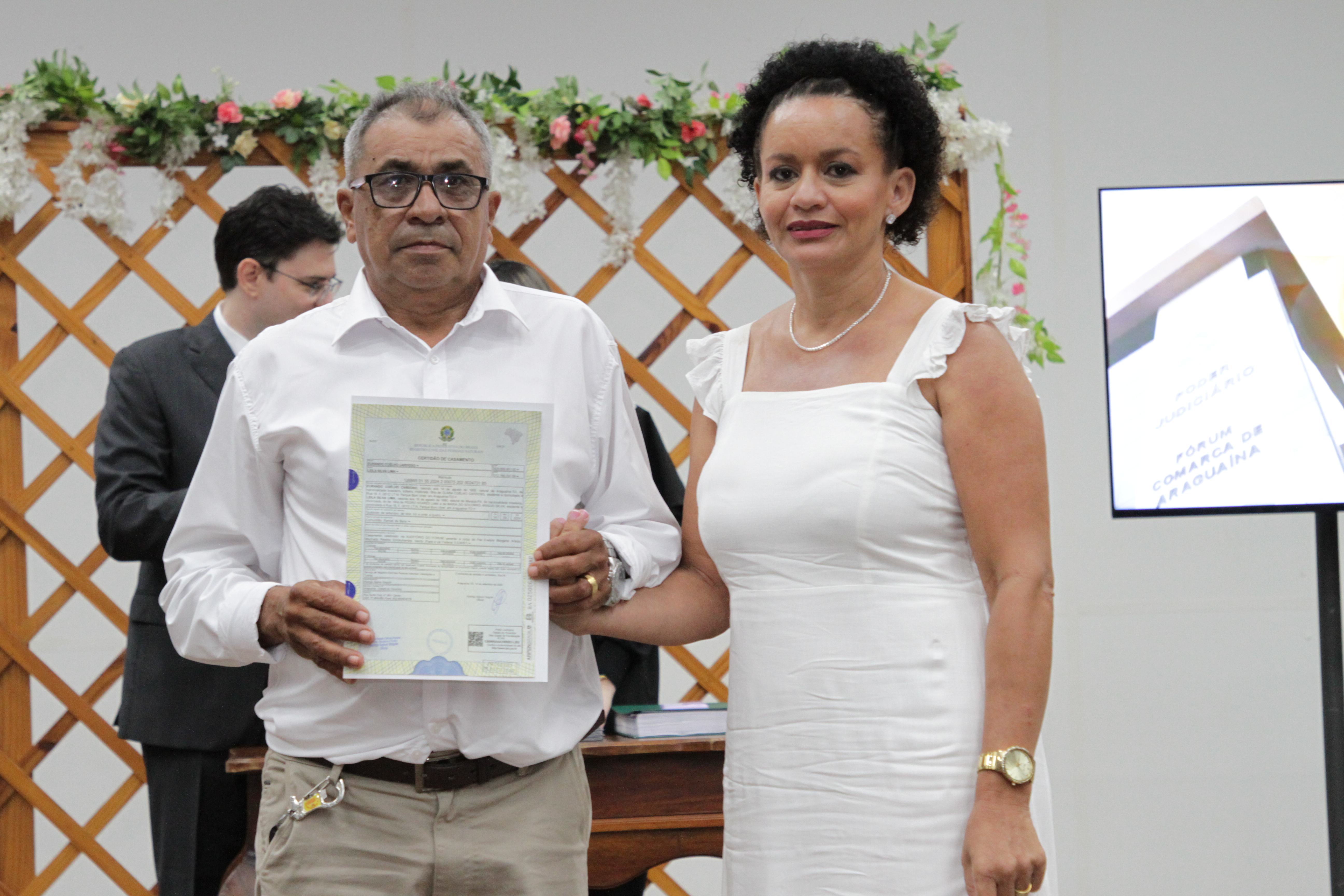 Fotografia colorida que mostra um casal heterossexual posando para a imagem segurando uma certidão de casamento. Ambos usam branco. 