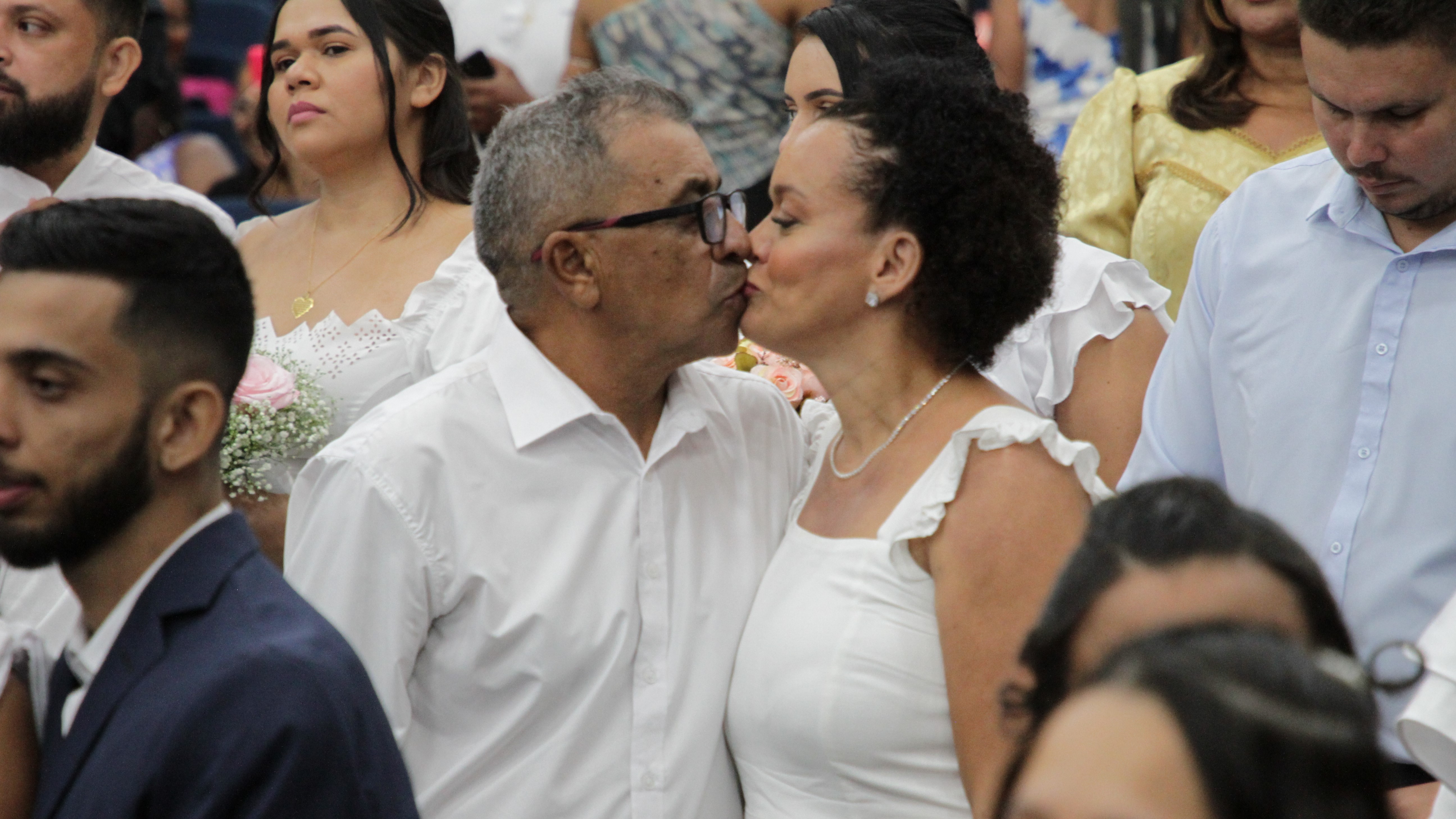 Fotografia colorida que mostra um casal heterossexual se beijando. Eles usam branco e há outros casais em volta
