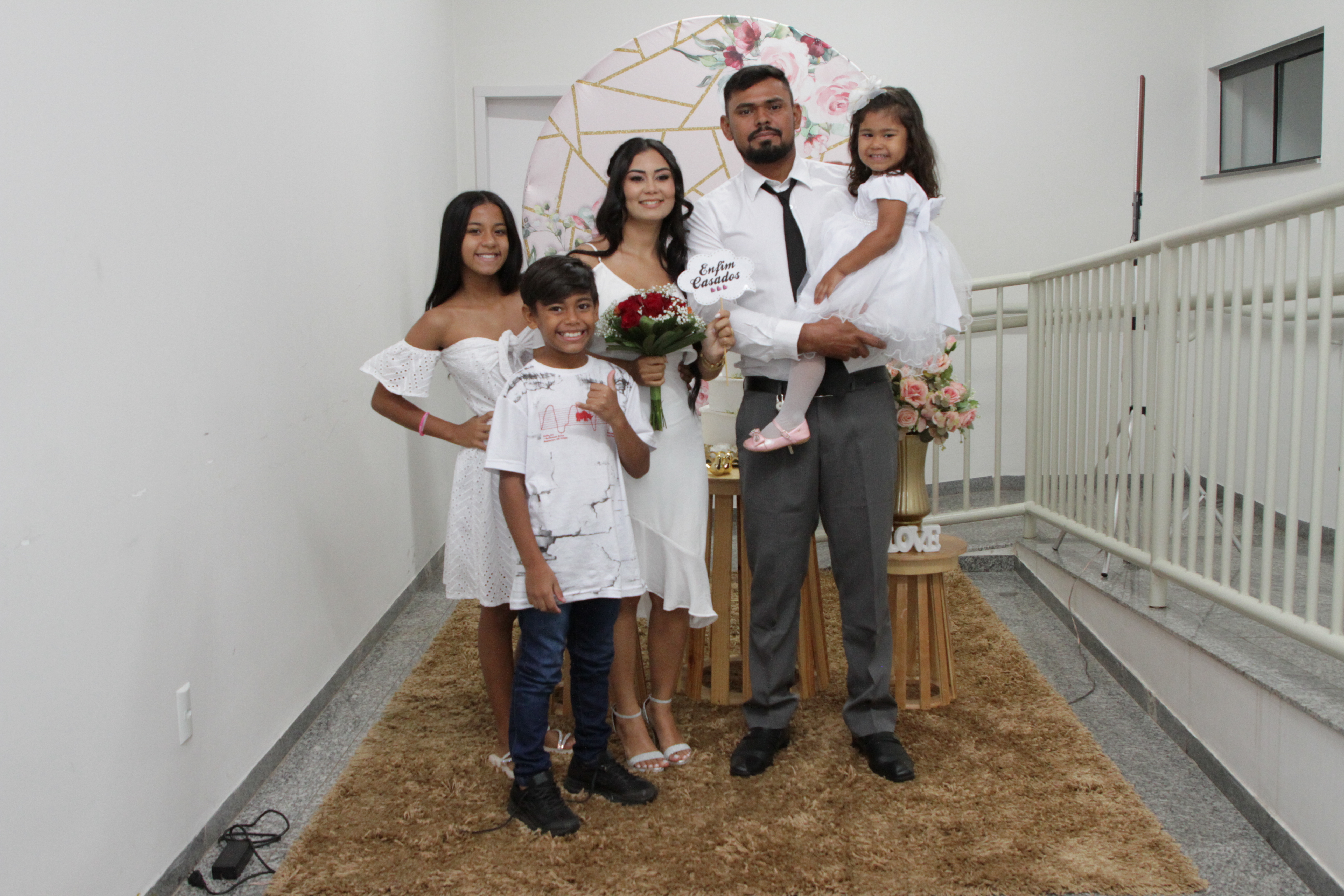 Imagem colorida que mostra um casal e três crianças (sendo uma delas um menino) posando para a foto.  O homem segura uma menina no colo e a mulher um buquê de rosas vermelhas e a placa : "Enfim casados"
