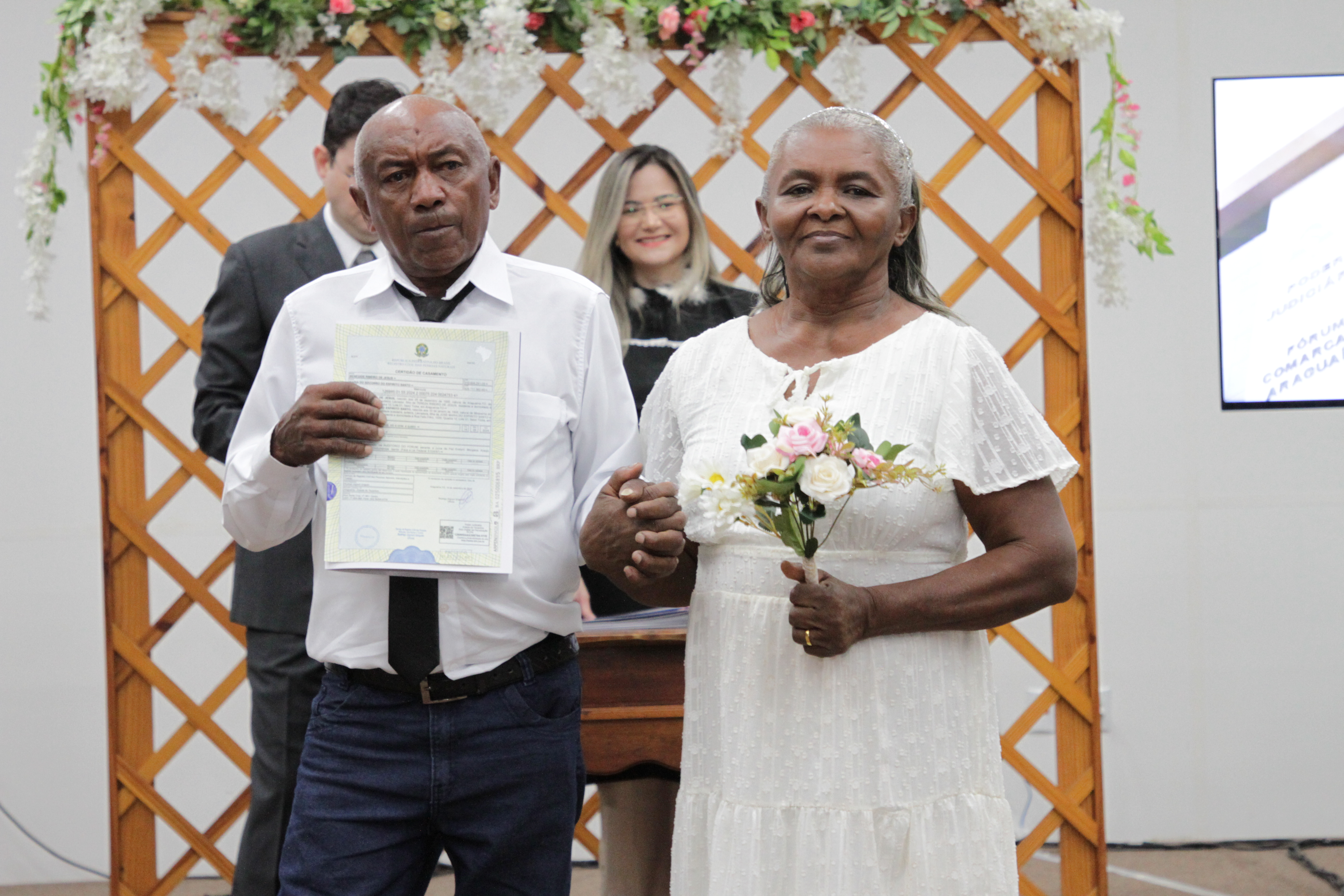 Fotografia colorida que mostra um casal heterossexual posando para a foto. A mulher segura um buquê de flores. 