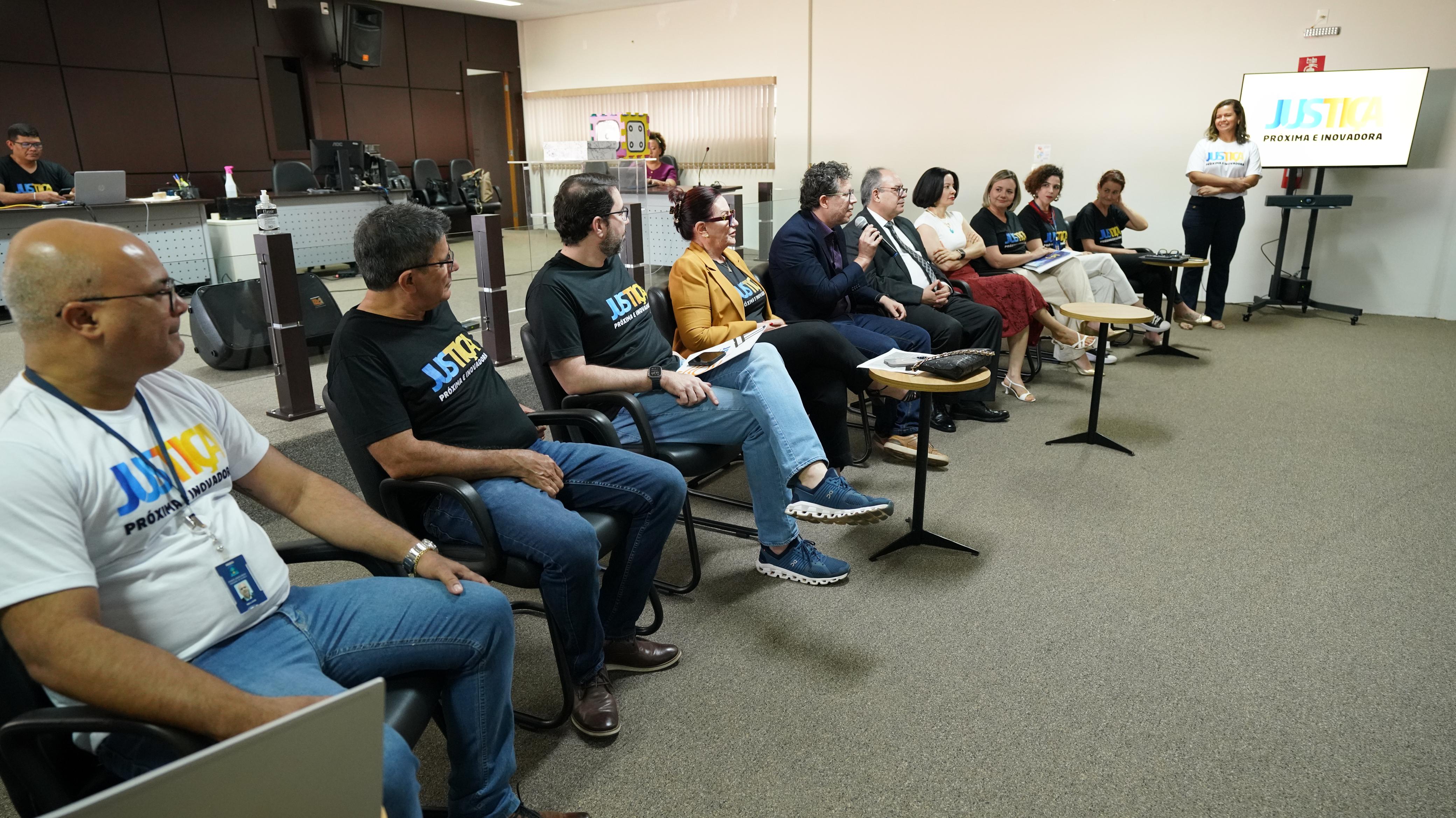 Fotografia colorida que mostra a diretora de Comunicação explicando algo para a plateia, em um auditório. Ao fundo, vê-se uma tela de televisor com o nome Justiça mais  próxima e inovadora