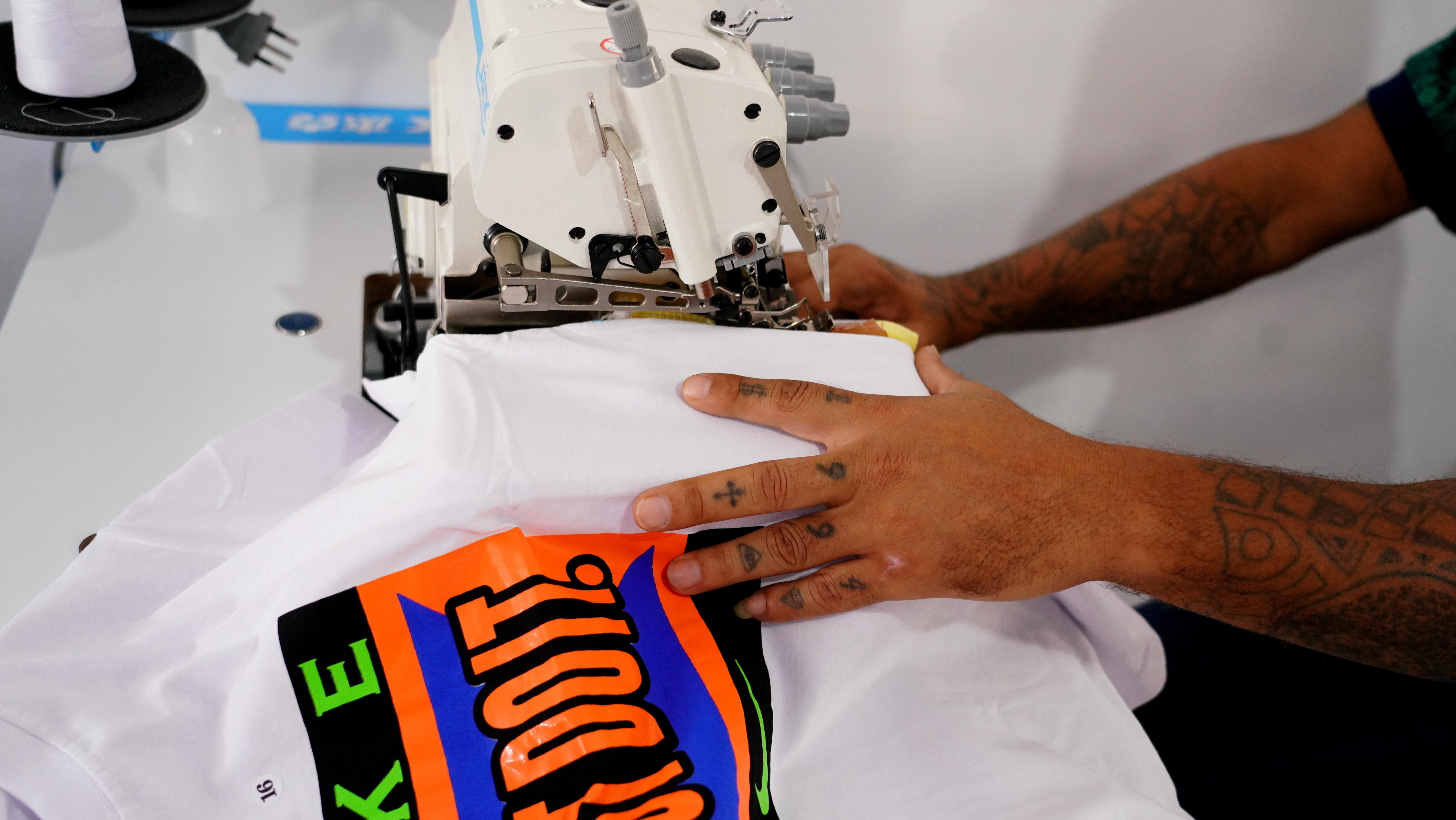 Fotografia colorida que mostra uma mão masculina, tatuada, costurando uma camiseta