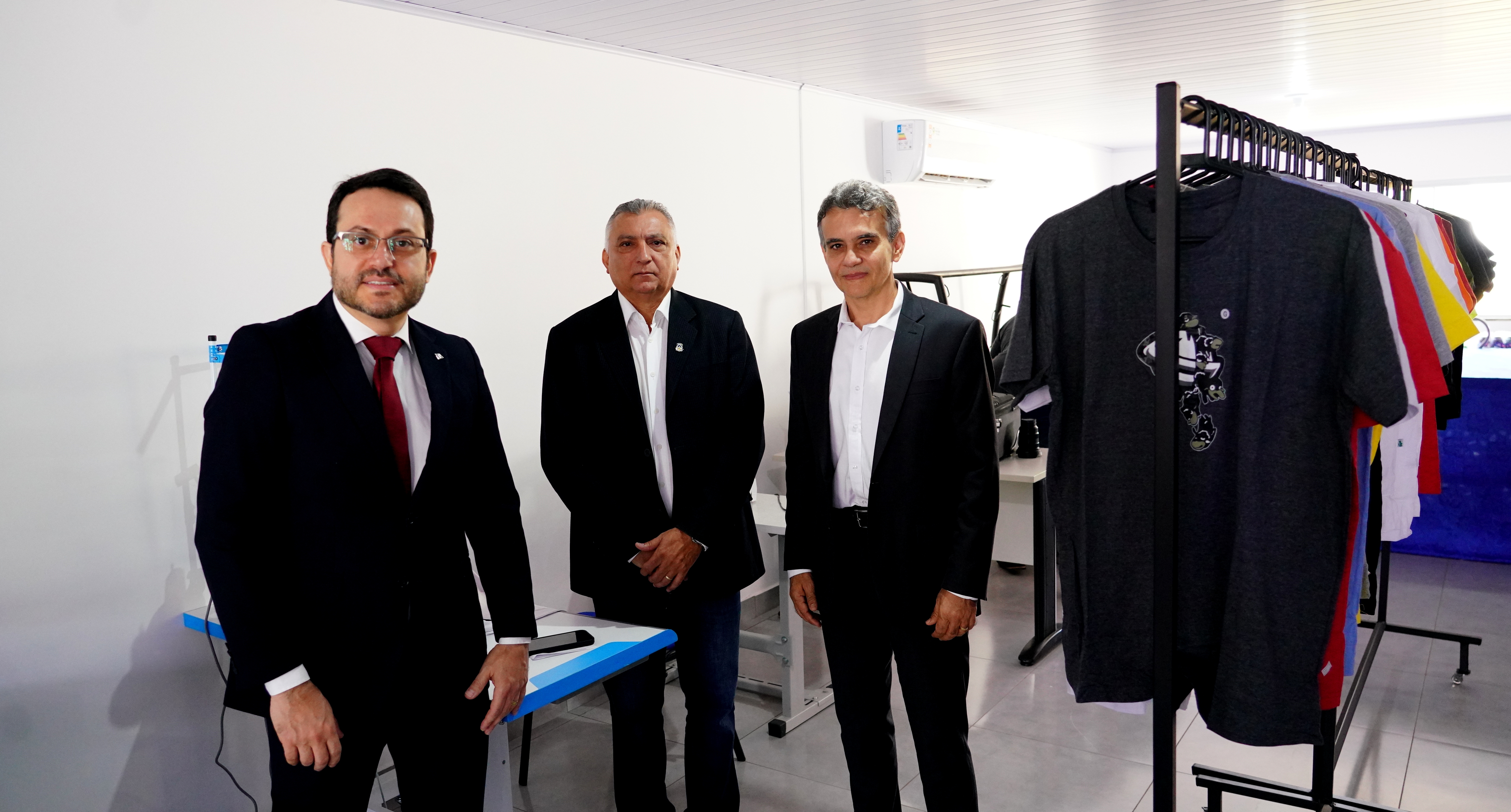 Fotografia colorida de três homens brancos, de terno e gravata posando pra fotos. Ao fundo, camisetas estão expostas. 
