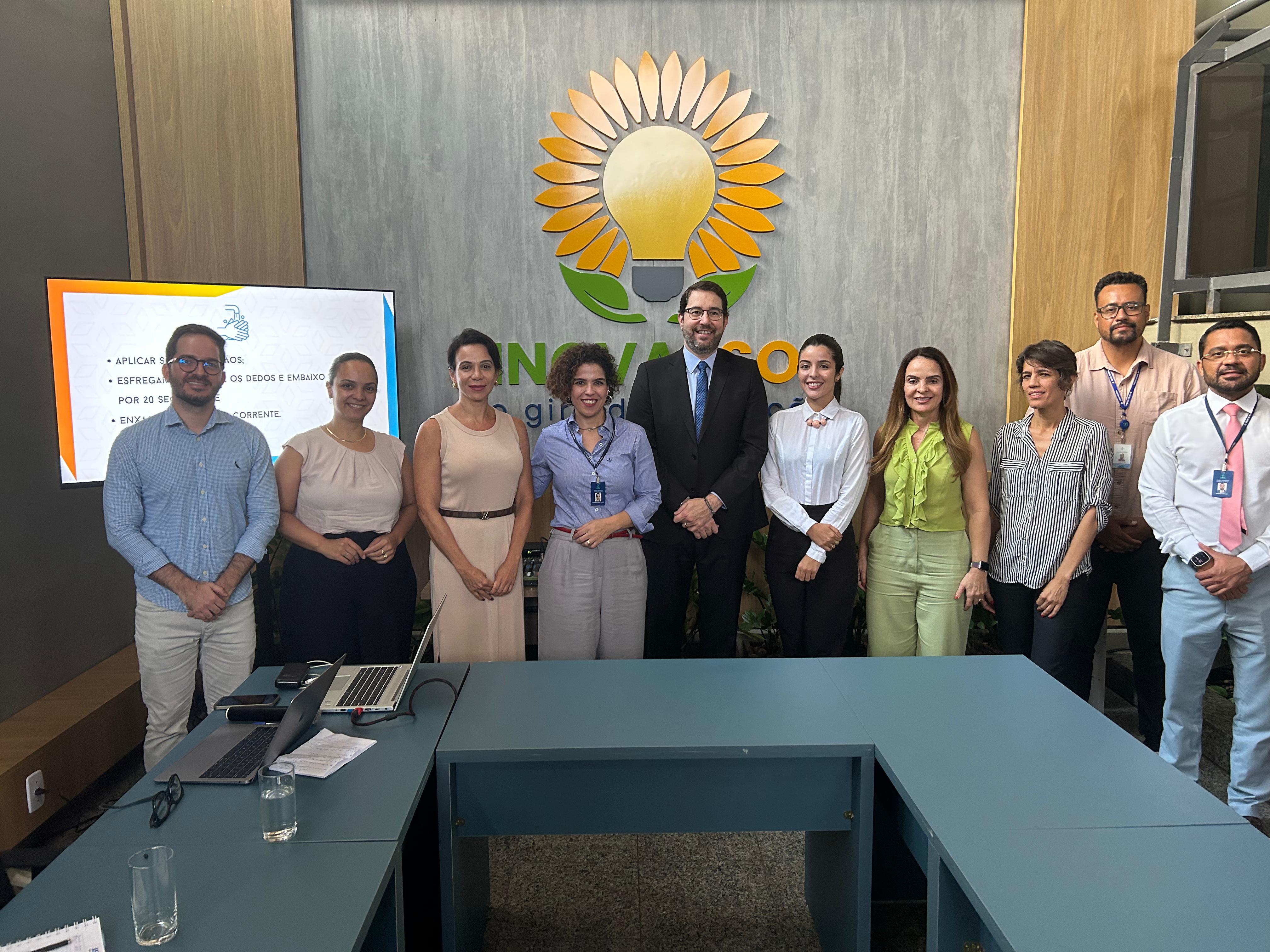 Pessoas com roupas coloridas estão em pé pousando para foto; à frente uma mesa azul e no fundo um painel com um flor de girassol