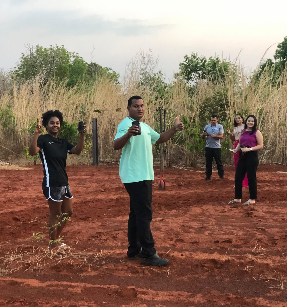 Cinco pessoas, sendo três do sexo masculino e duas do feminino em pé, segurando mudas de árvore em uma área verde