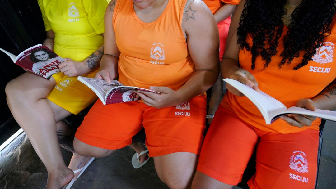 três reeducandas de costa, com uniforme amarelo, lendo livros