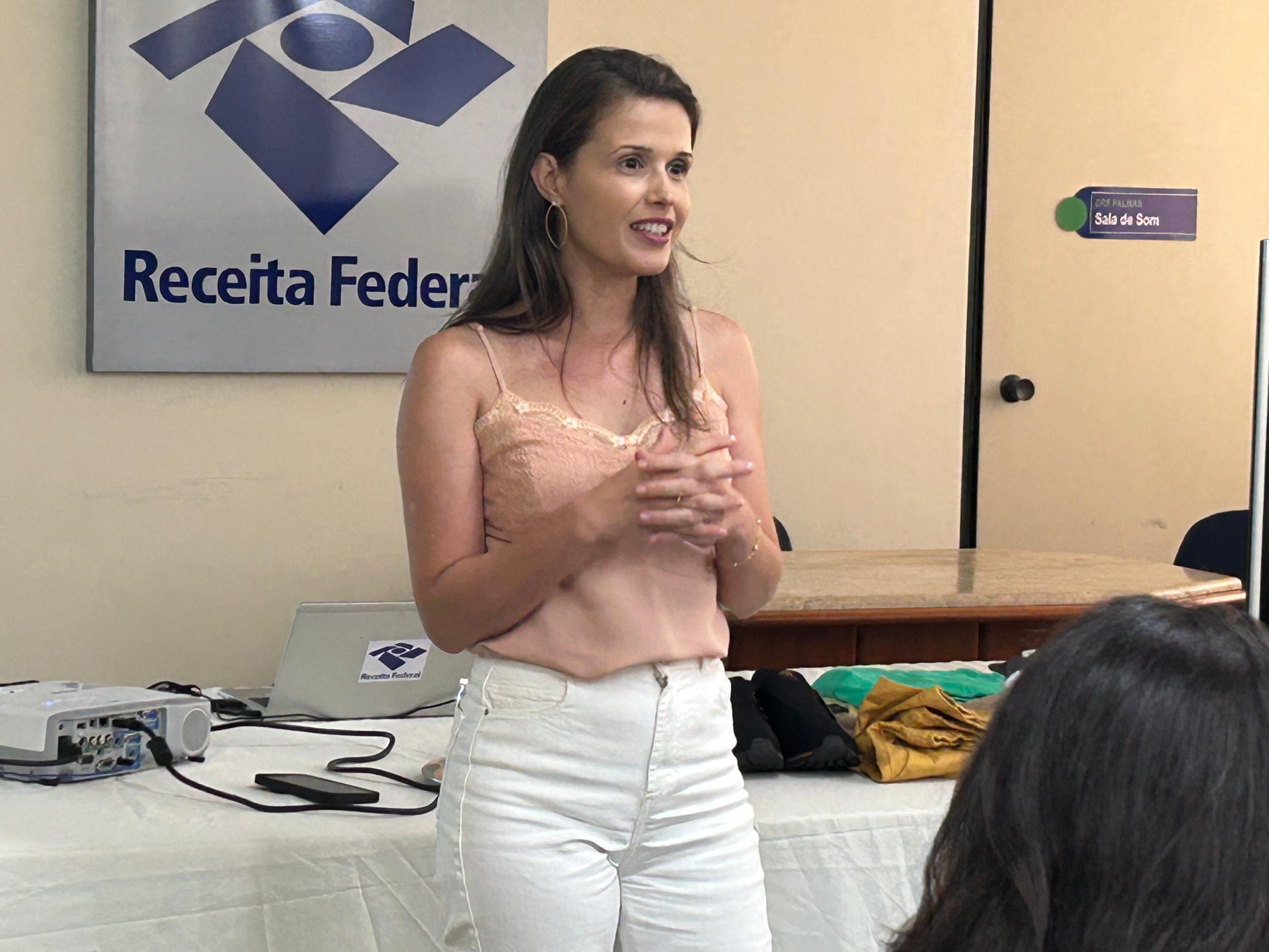 Francine de Paula, uma mulher jovem, de cabelos escuros e longos, fala; ela usa calça branca e blusa rosa; ao fundo uma placa com a logo da Receita Federal