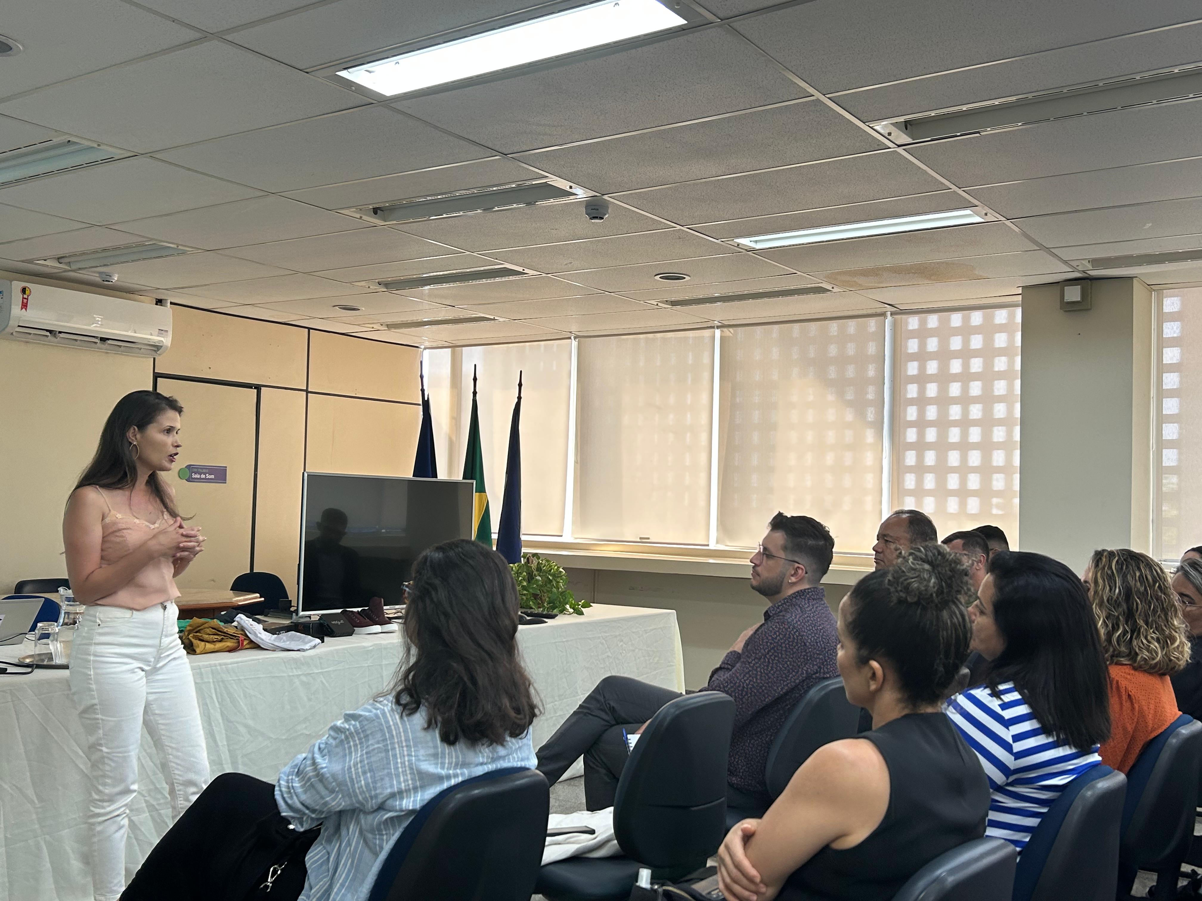 Em uma sala, algumas pessoas sentas de costa, à frente Francine fala em pé; ela é uma mulher jovem de cabelos grandes e escuros, usa calça branca e blusa rosa