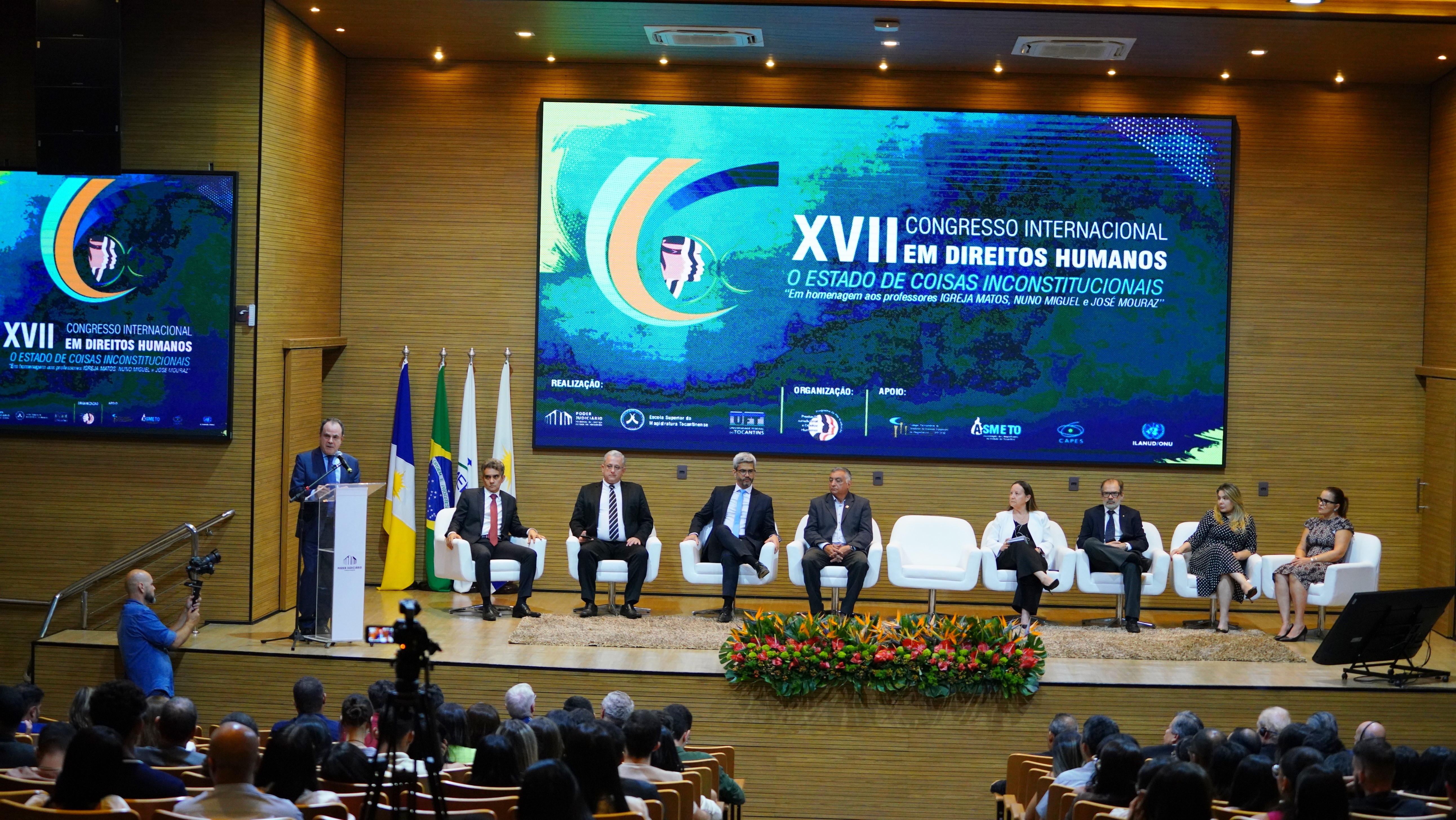 Fotografia colorida mostra o desembargador Marco Villas Boas falando no púlpito e sendo observado por autoridades no palco e na plateia na abertura do evento 