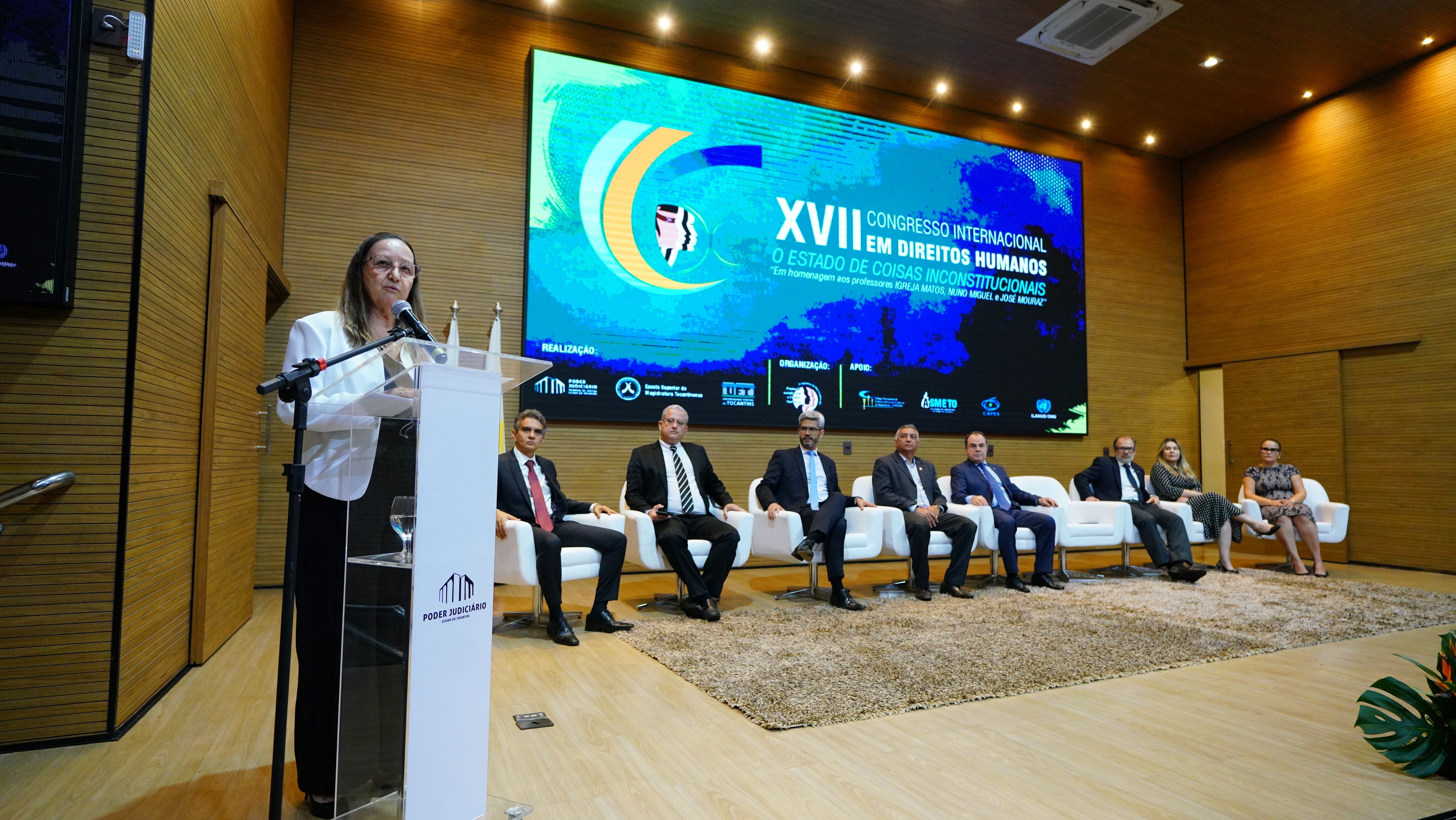 Imagem colorida que mostra uma mulher branca, de óculos e usando terno branco e calça preta falando de um púlpito observada por homens e mulheres no palco. Ao fundo tem um telão com o tema do Congresso.