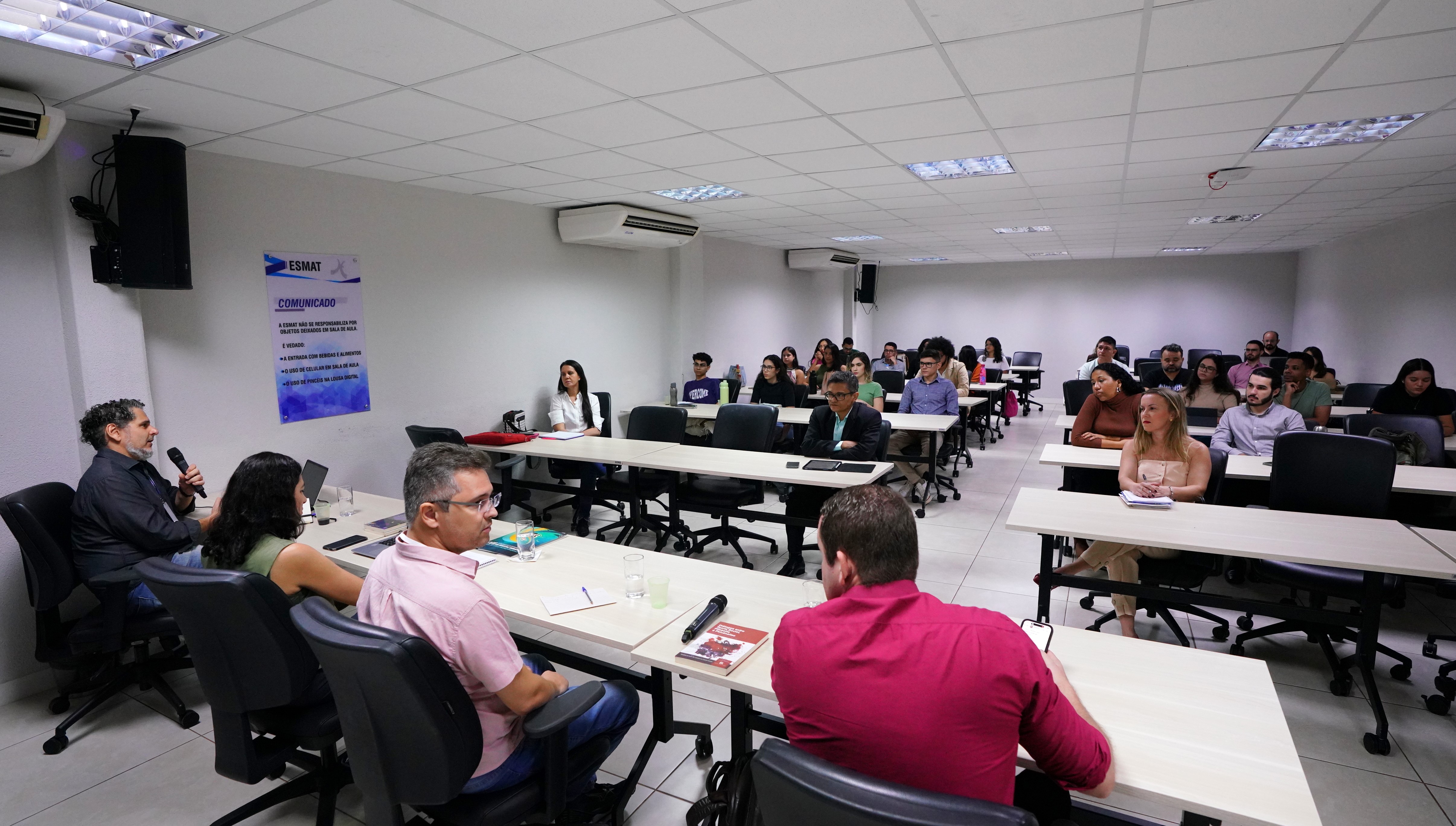 Fotografia colorida que mostra quatro pessoas sentadas em volta de uma mesa discursando para uma plateia