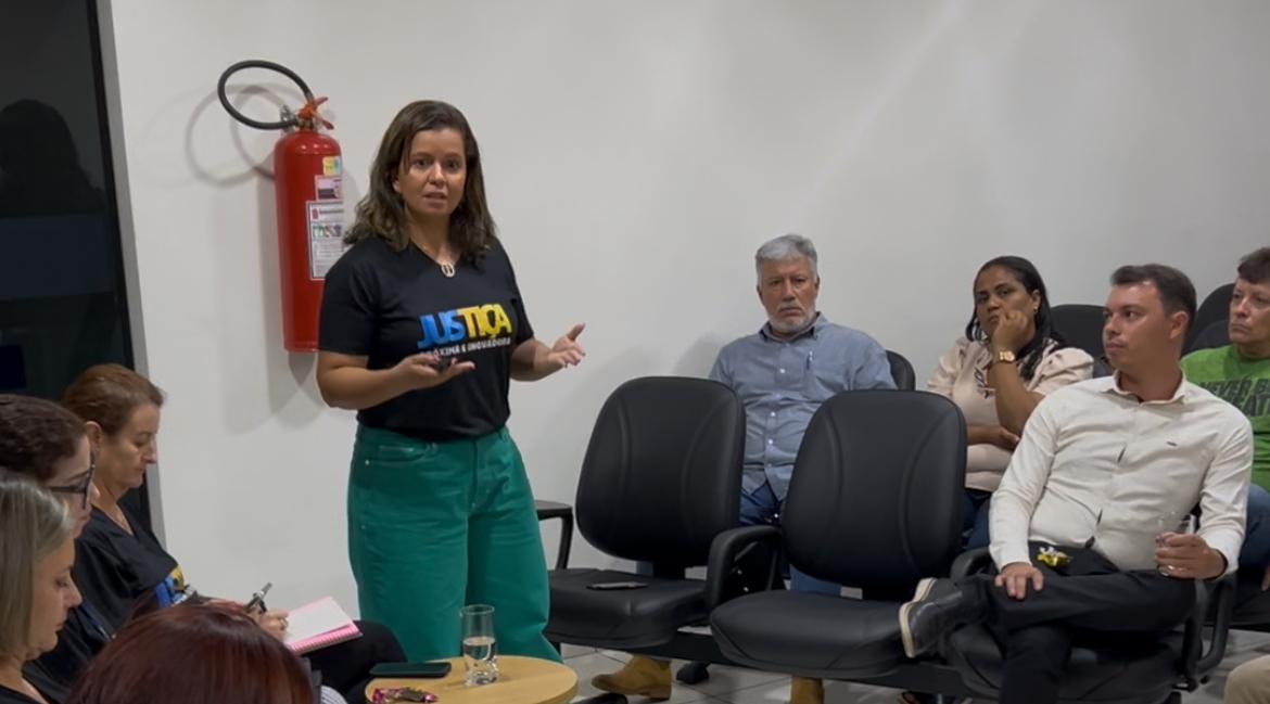 Imagem colorida que mostra uma mulher negra,de cabelos lisos, usando uma calça verde e camiseta preta escrita: Justiça mais próxima e inovadora. Ela fala e é observada pelas pessoas em uma sala.