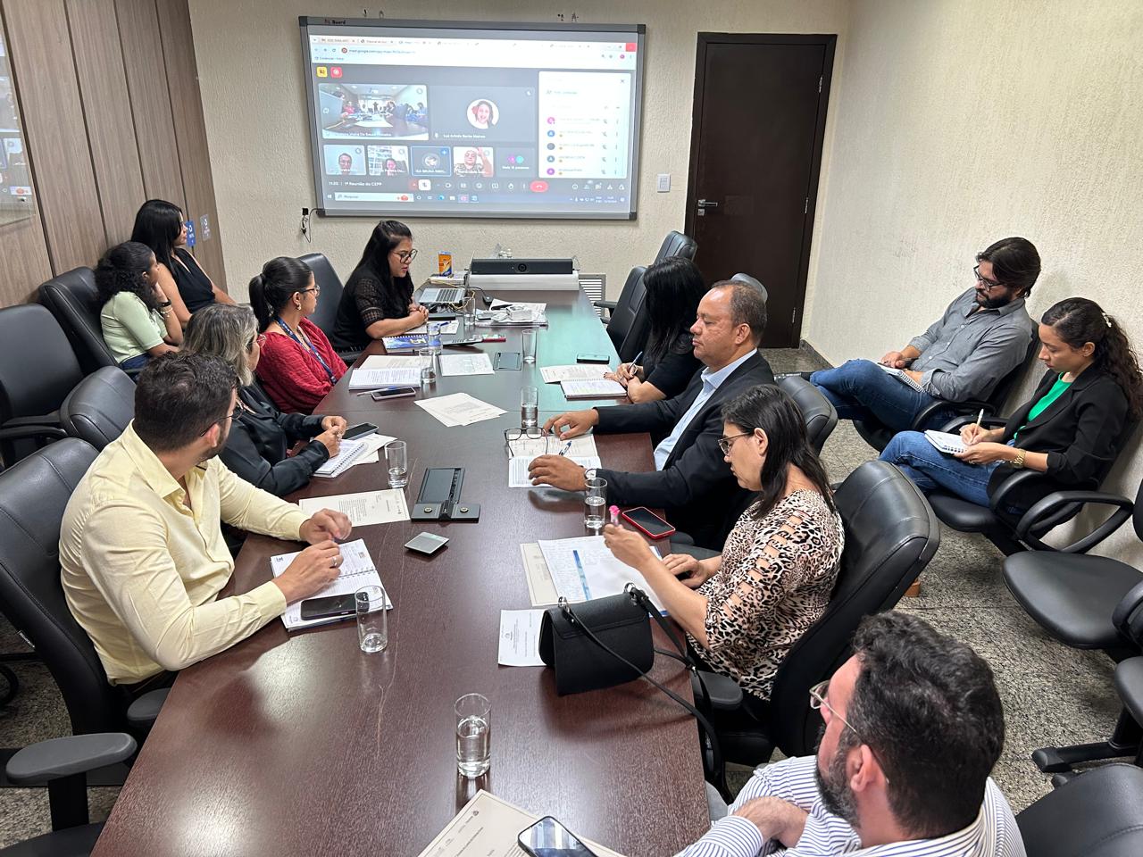 Imagem em uma sala de reunião; pessoas sentadas em uma mesa de madeira e à frente um telão com uma reunião virtual