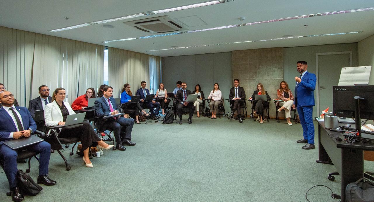 Imagem mostra um homem com terno azul e em uma roda pessoas sentadas em uma sala
