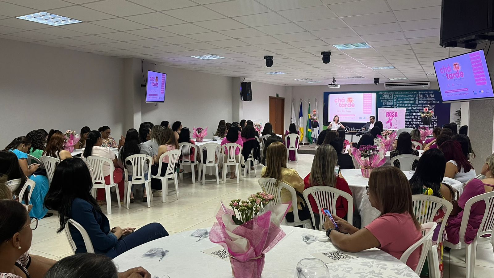 Fotografia colorida que mostra uma sala repleta de mulheres usando rosa assistindo a uma palestra. Nos televisores se vê o nome Chá da Tarde e o simbolo do Outubro Rosa