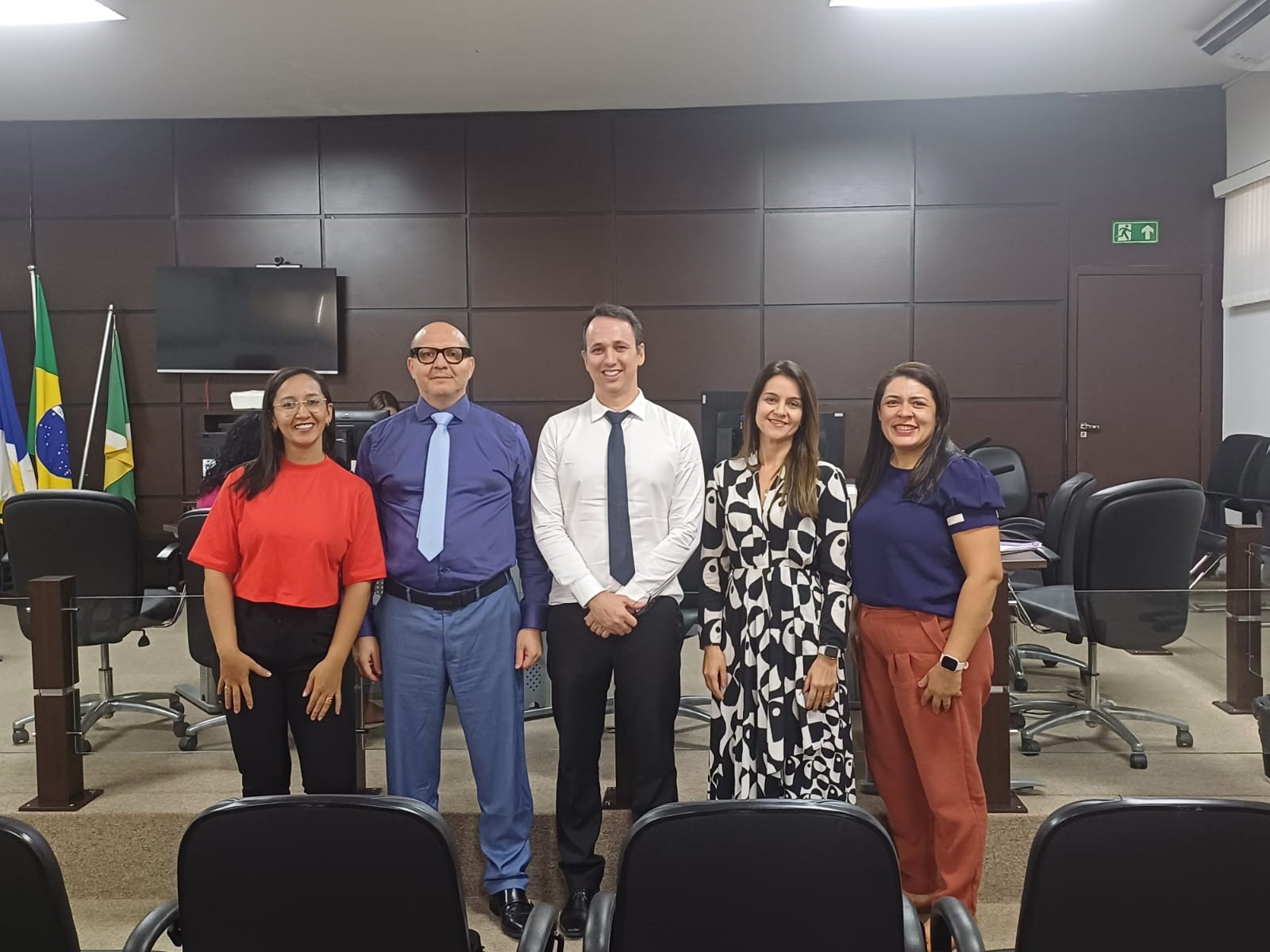 Três mulheres brancas e dois homens brancos, com roupas coloridas estão em pé posando para foto