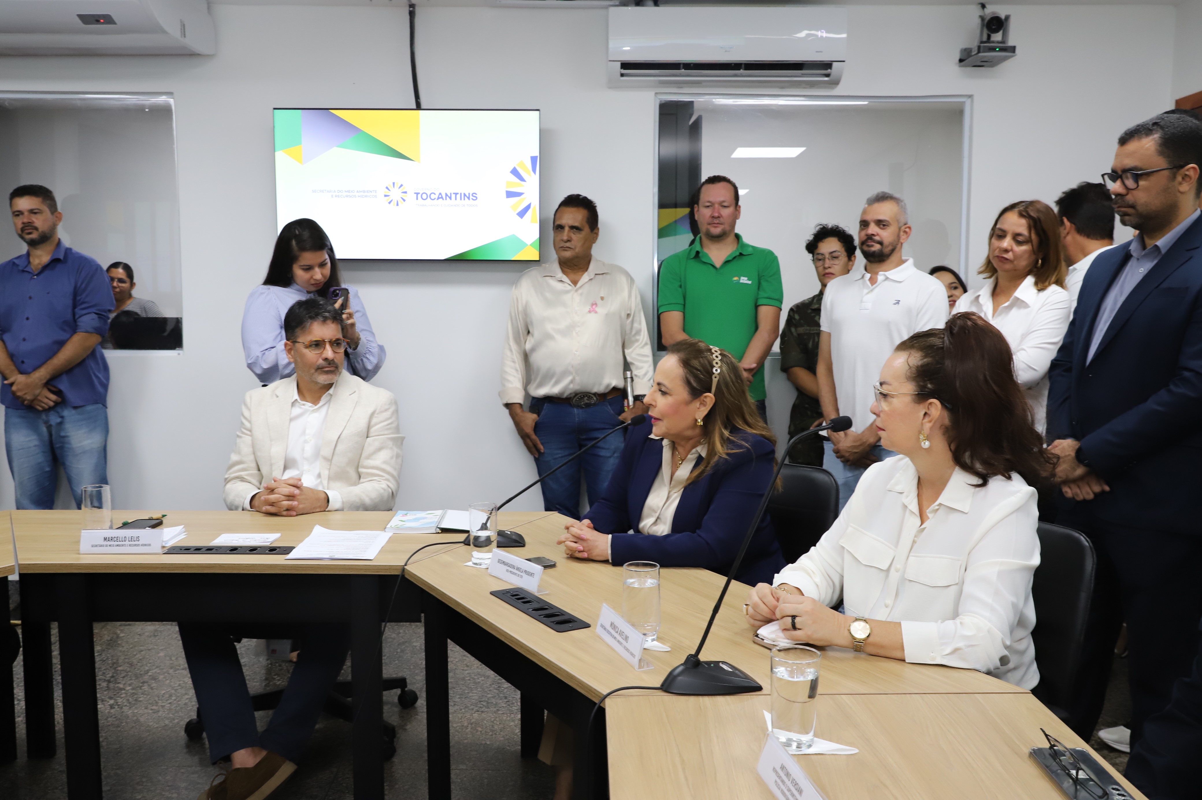 Fotografia colorida que mostra autoridades (homens e mulheres) sentados numa mesa e outras pessoas observam