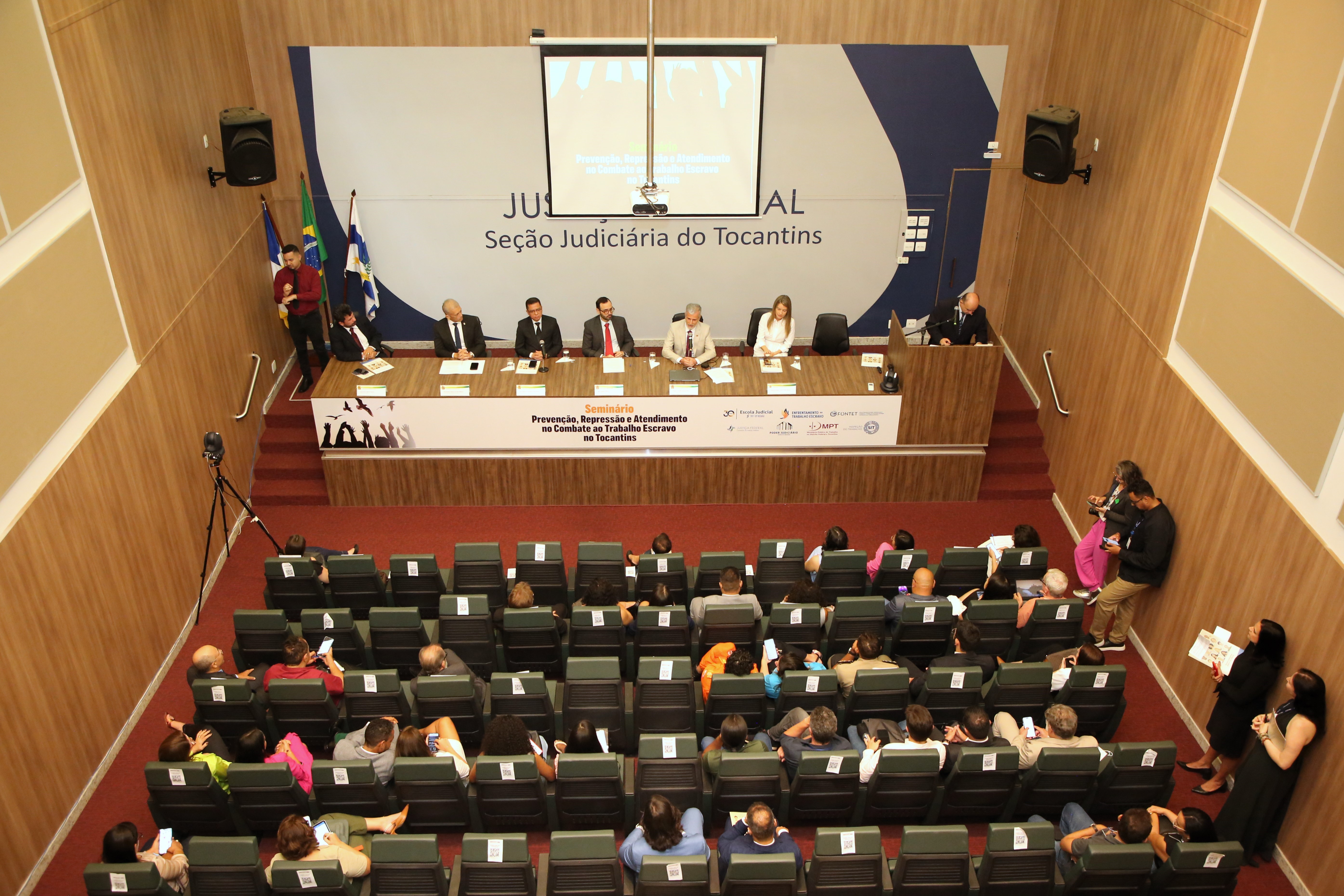 Fotografia colorida que mostra um auditório cheio de pessoas e autoridades em uma mesa e frente ao palco