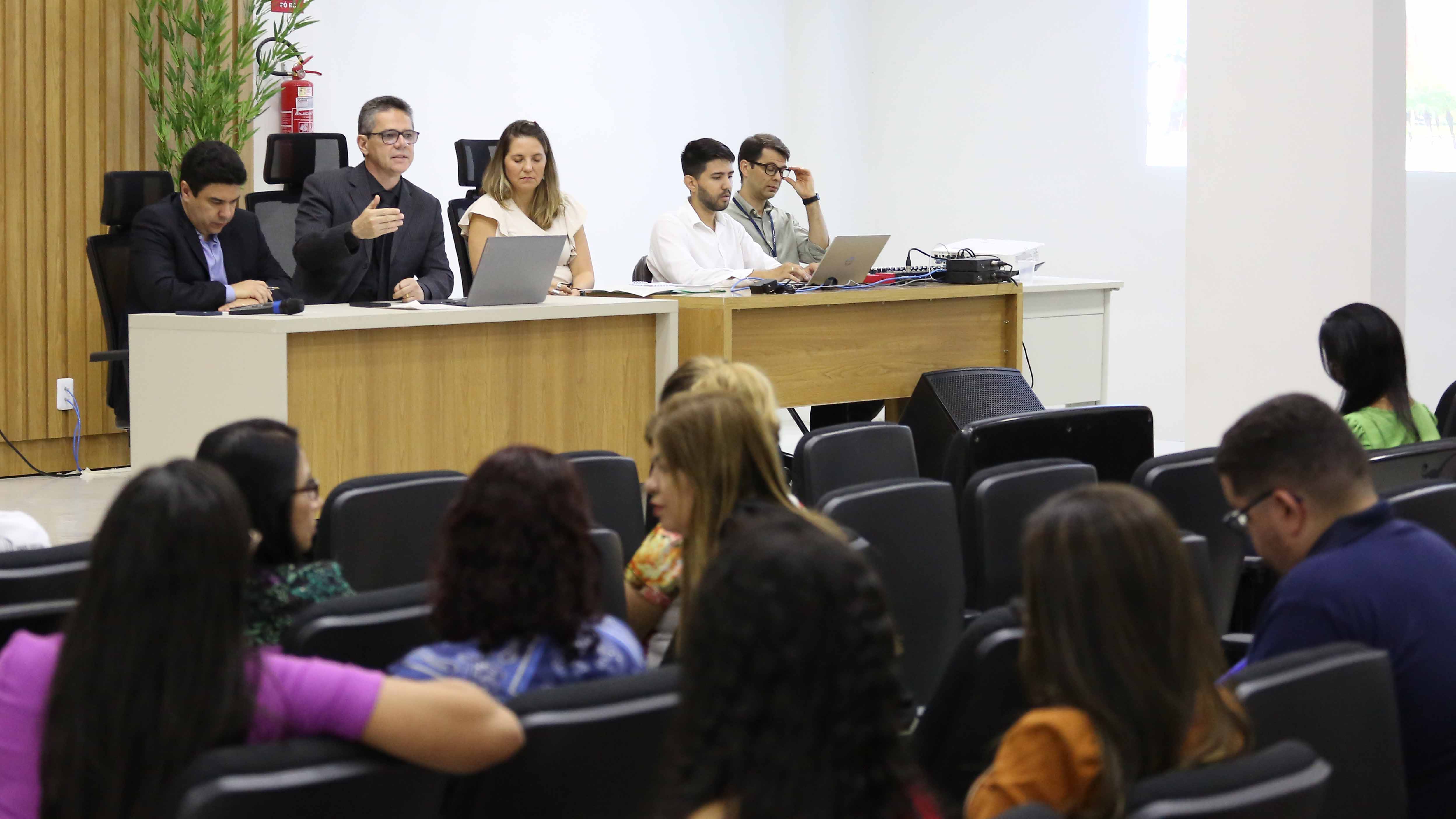 Homens e mulheres sentados no auditório assistindo um homem de terno preto sentado falando e gesticulando com as mãos. Ele usa óculos de grau e está sentado à uma mesa de madeira cor clara, ladeado por outras pessoas