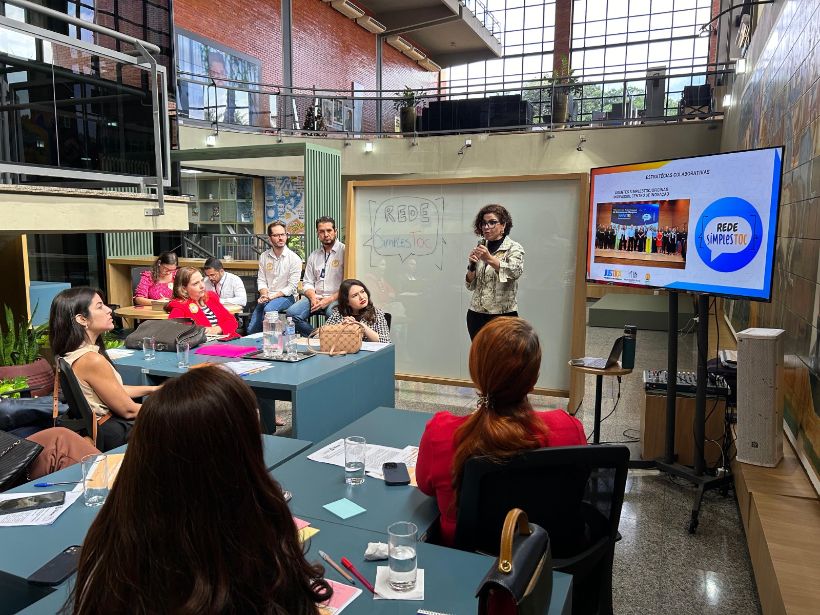 Fotografia colorida que mostra uma mulher branca de cabelos cacheados falando pra uma plateia formada por homens e mulheres no Inovassol