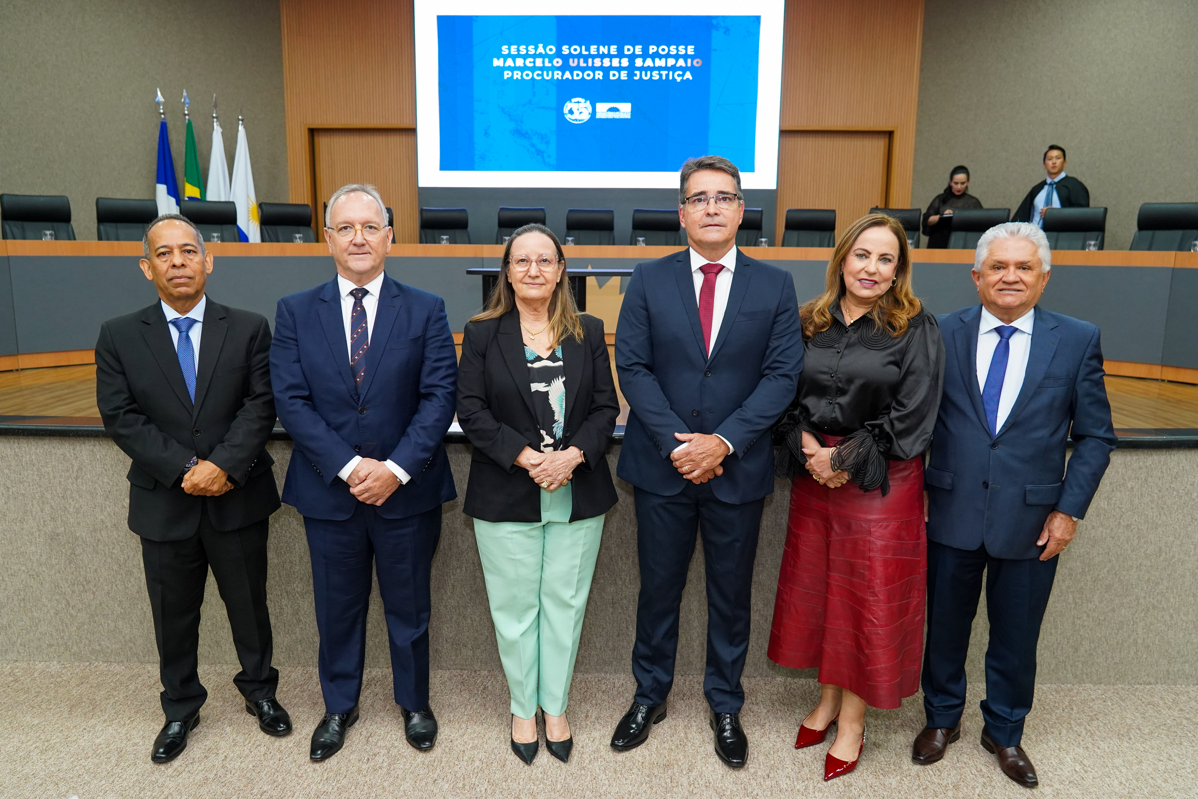 Imagem com quatro homens e duas mulheres em pé, usando roupas coloridas e ternos, pousando para a foto
