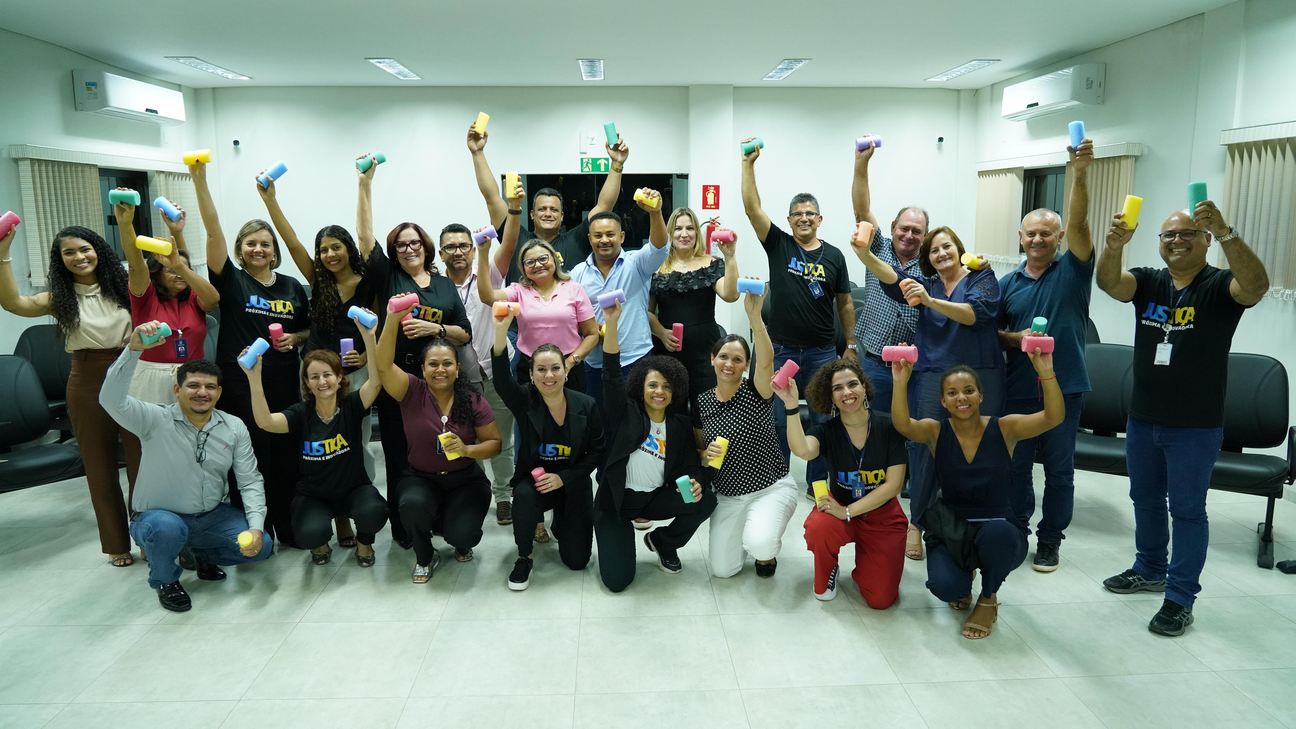 A imagem tem um grande grupo de pessoas sorrindo que está reunido em uma sala, cada uma segurando um pequeno objeto colorido acima da cabeça. Muitas pessoas estão usando camisetas com a palavra "Justiça" impressa nelas.