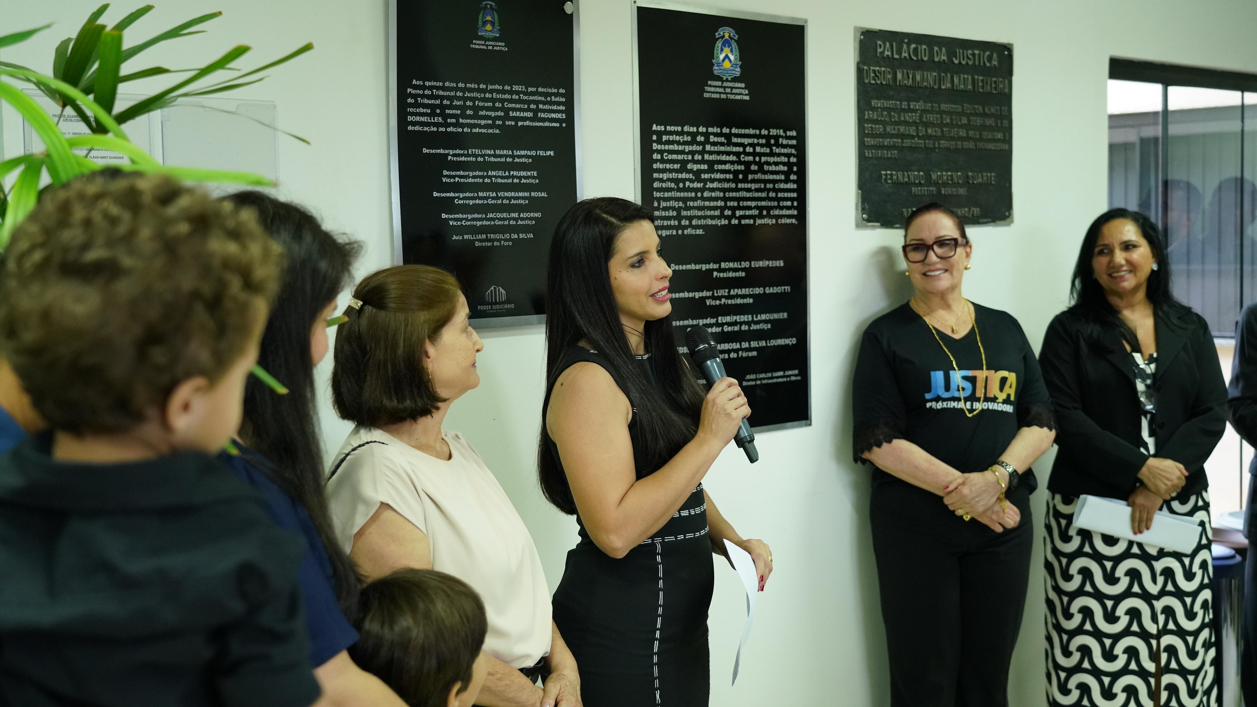 Mulher de roupa preta está em pé segurando um microfone ela fala para várias pessoas ao redor