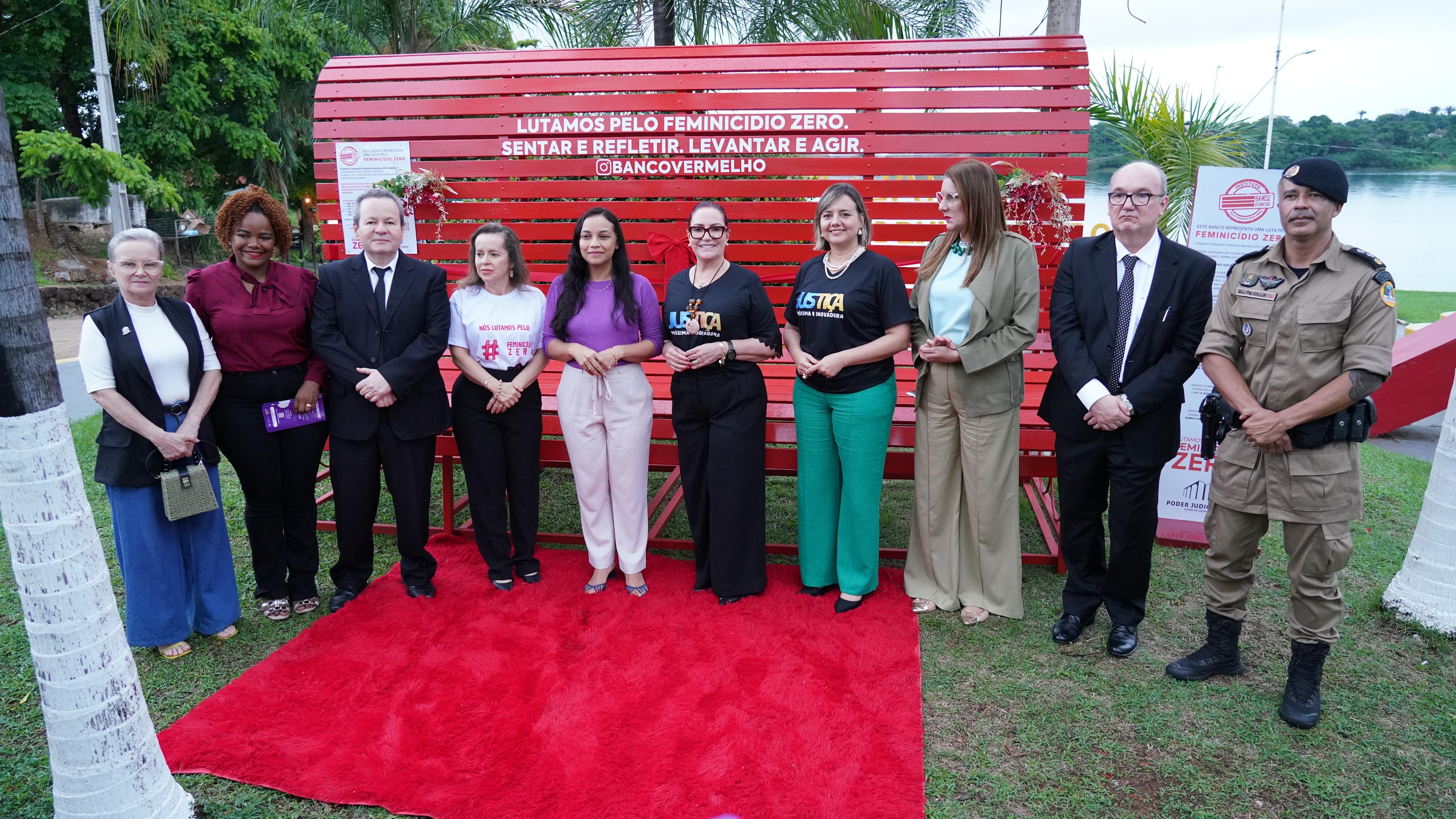 Homens e mulheres e pé sobre um tapete vermelho em cima de um gramado onde está instalado um banco vermelho gigante