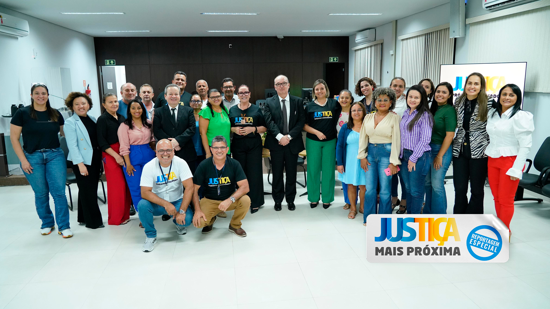 Homens e mulheres posam para foto em sala de paredes brancas e persianas beges. Ao fundo, uma parede com acabamento em madeira escura