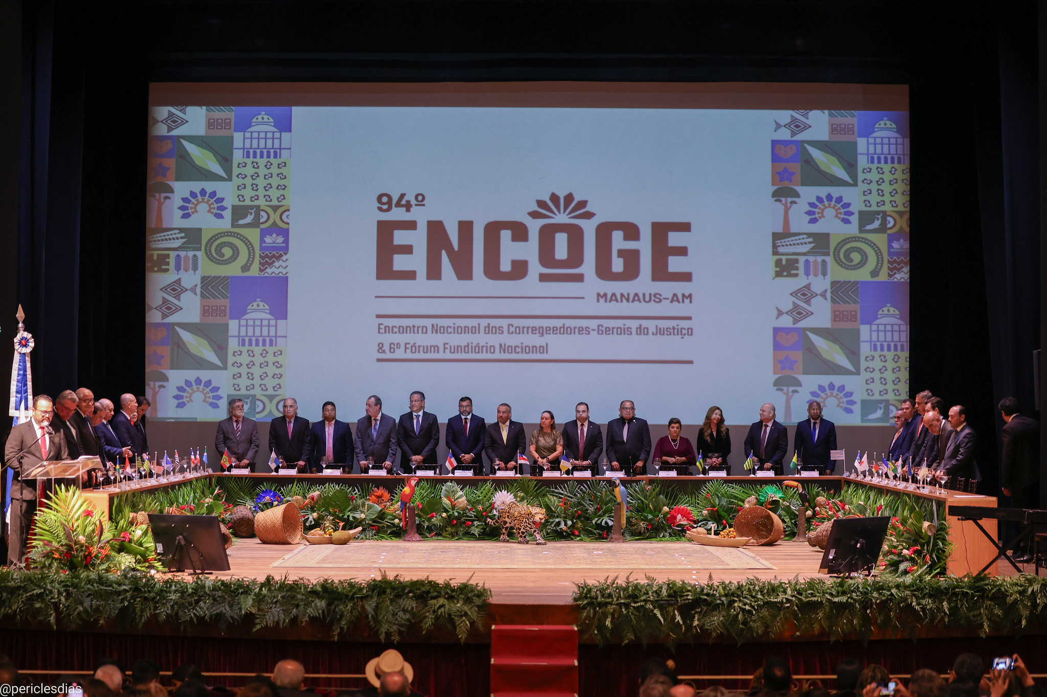 Fotografia colorida onde um grande grupo de pessoas estão distribuídos em volta de uma mesa retangular aberta. Nós pés da mesa, estão vários adornos de plantas e frutas nativas por todas a sua extensão e ao fundo um grande telão mostra uma imagem com bordas com padrões de imagens diversos e ao centro os dizeres  "94.º Encontro Nacional dos Corregedores-Gerais de Justiça (Encoge)" 