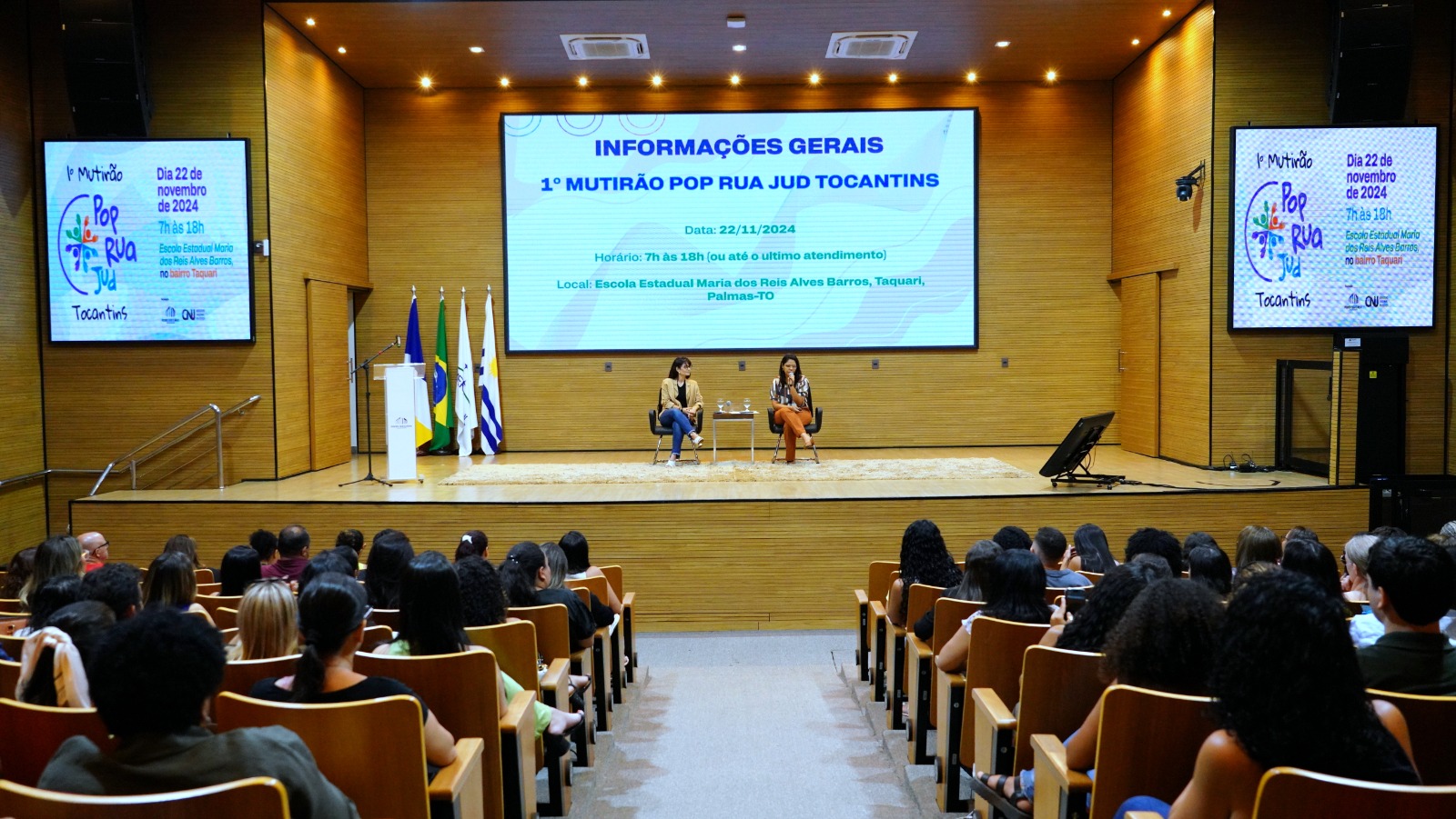 foto colorida horizontal mostra o auditorio do tjto com pessoas sentadas na plateia. No palco duas mulheres estao sentadas em duas cadeiras, uma delas fala ao microfone, no telão informações sobre o pop rua jud