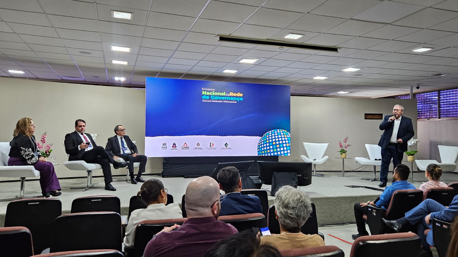 Pessoas estão sentadas em cadeiras vermelhas e outras em um palco falam ao público