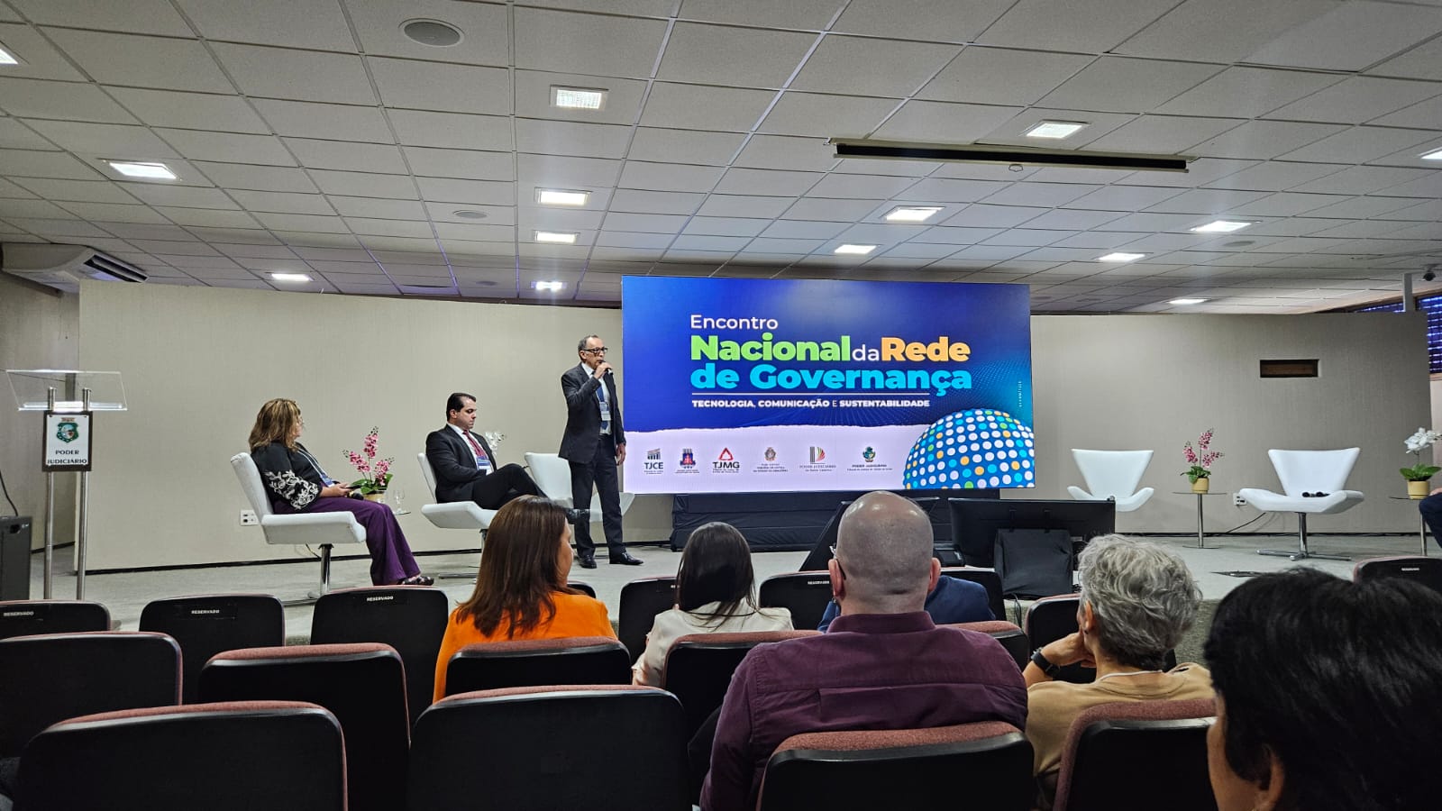 Foto mostra um lugar grande com paredes claras e cadeiras vermelhas; ao palco um homem em pé outro sentado e uma mulher sentada;  ao fundo um telão