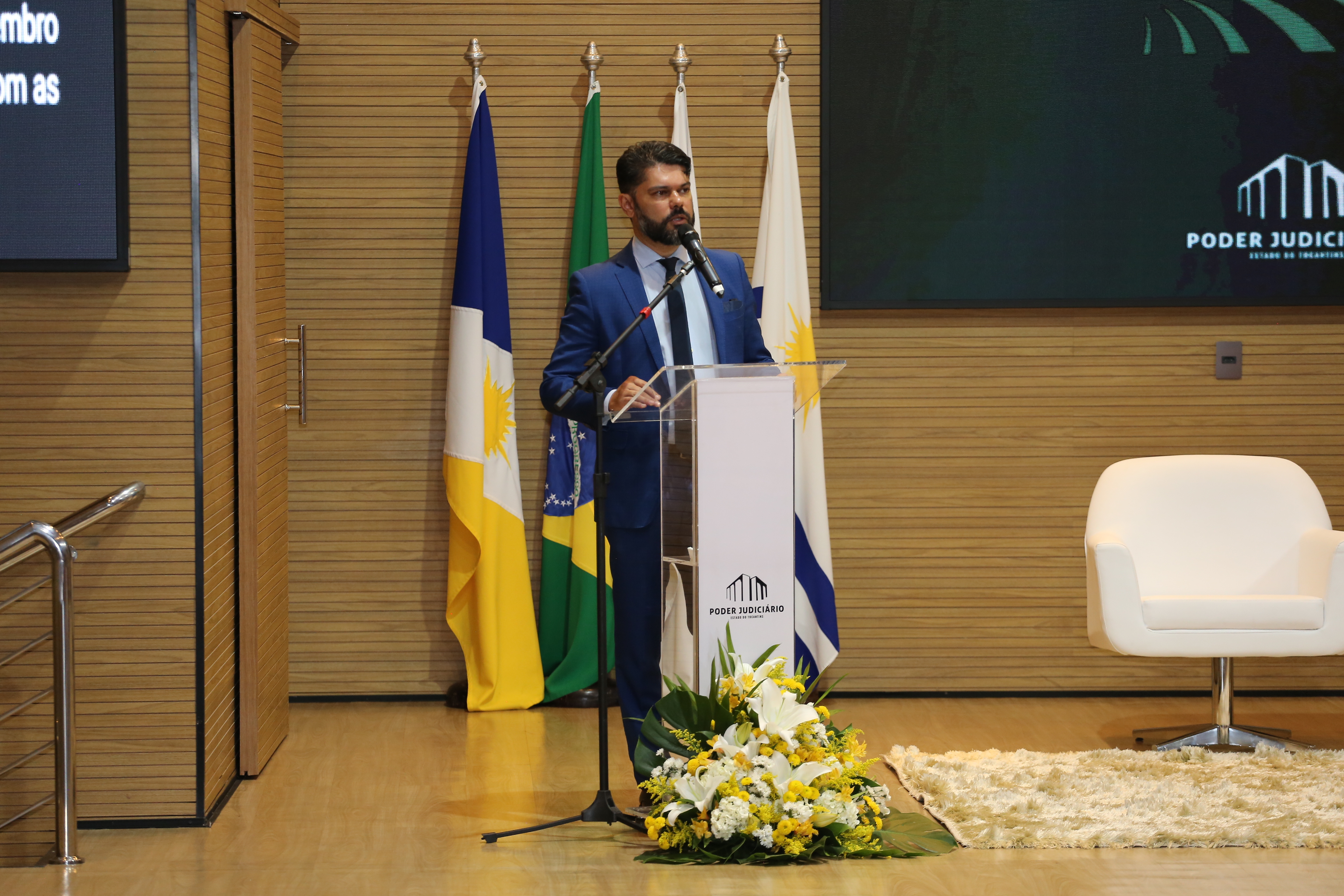 Fotografia colorida que mostra o juiz Wellington, um homem moreno, de barba, usando terno falando ao público do evento. 