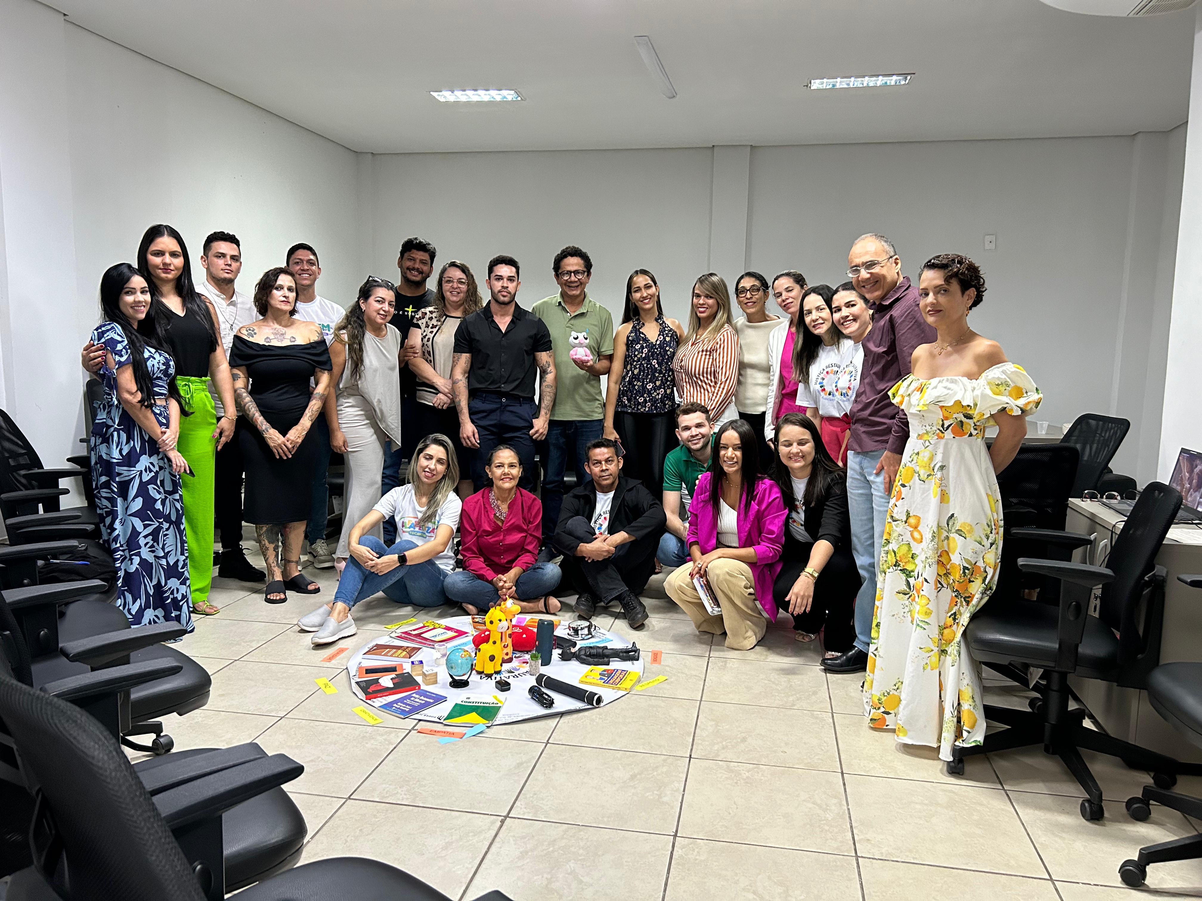 Fotografia colorida onde várias pessoas estão lado a lado posando para uma foto em uma sala branca. Em frente ao grupo, se encontra um circulo que simboliza uma dinâmica do projeto com alguns brinquedos em cima 